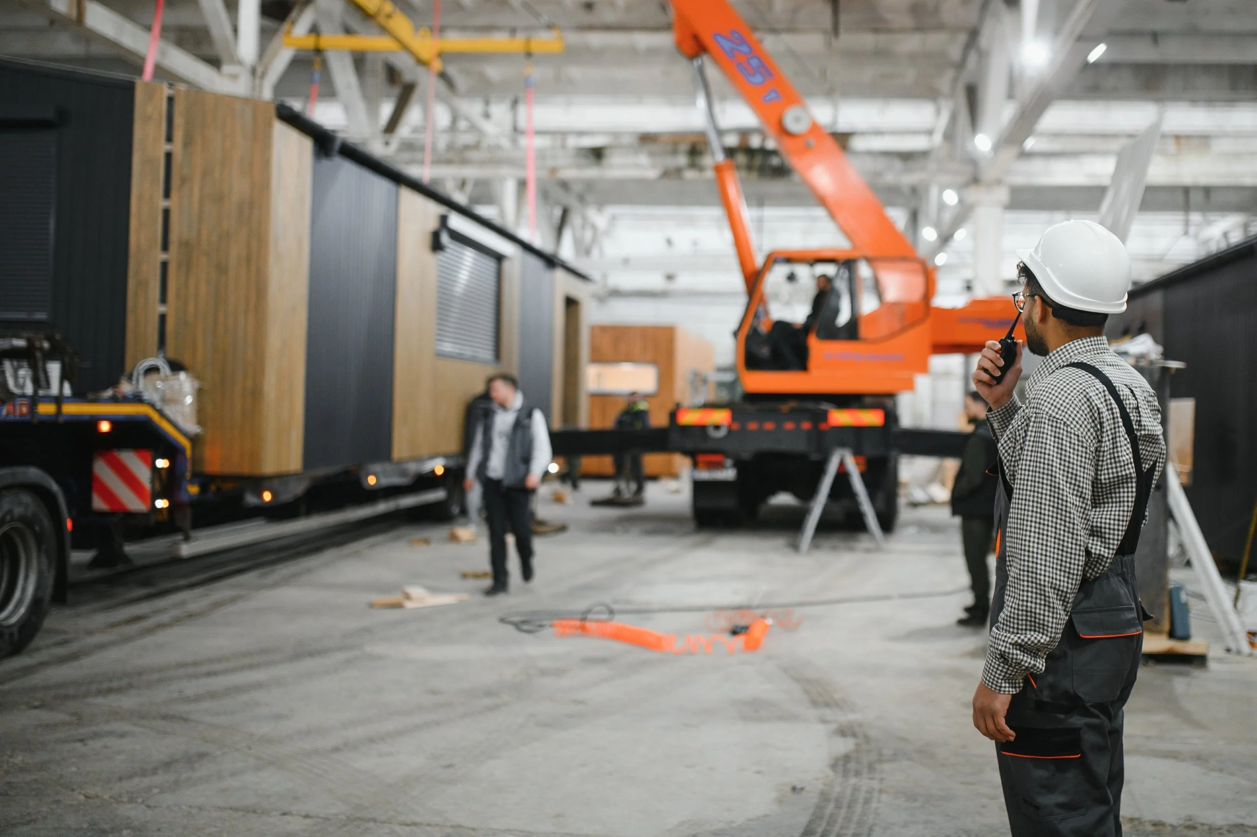 Construction workers inside a building with a large orange crane lifting materials, while one worker wearing a helmet and checkered shirt communicates via walkie-talkie.