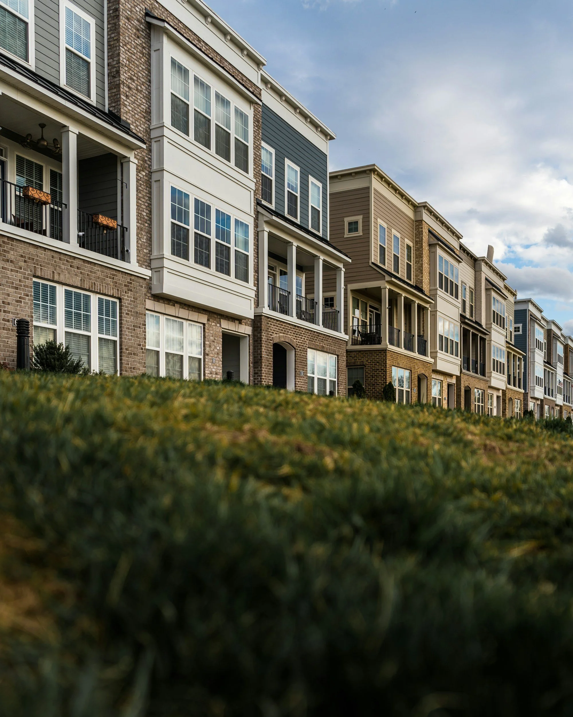 Row of multi-story residential apartments with brick and siding exteriors, balconies, and large windows, viewed from a grassy area.