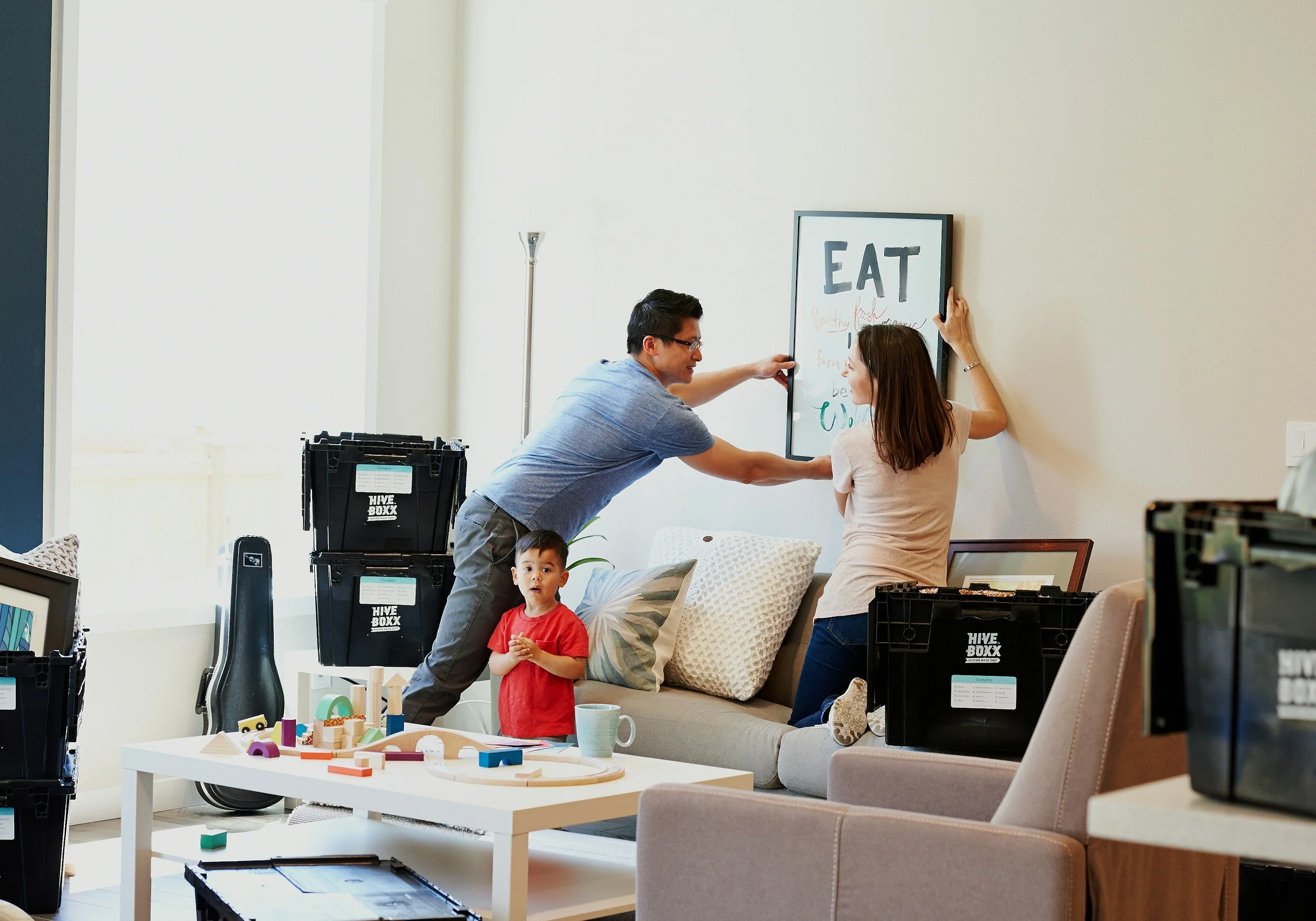A family rearranging a framed picture on the wall in a living room, with two children present and moving boxes around.