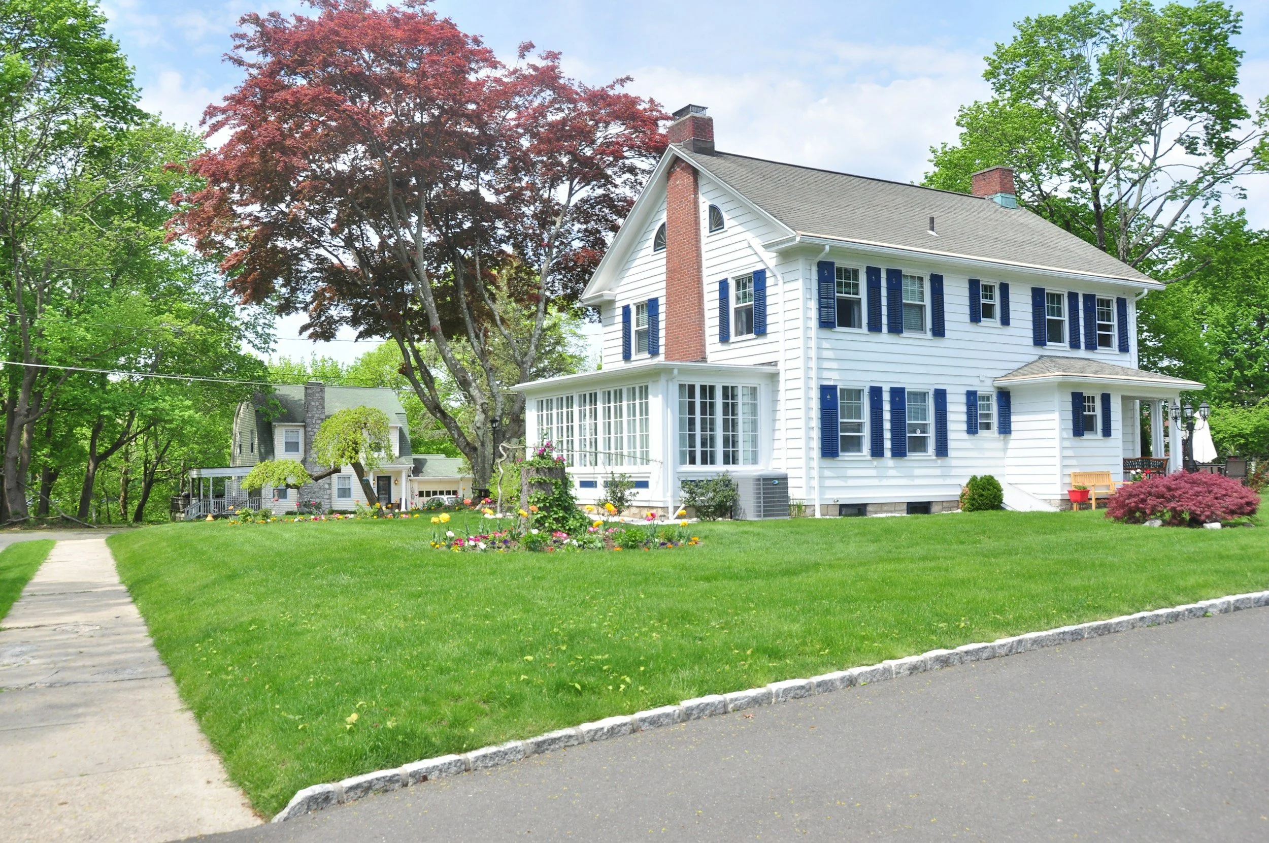 A white house with dark blue shutters and a brick chimney in a green yard with trees and a sidewalk.