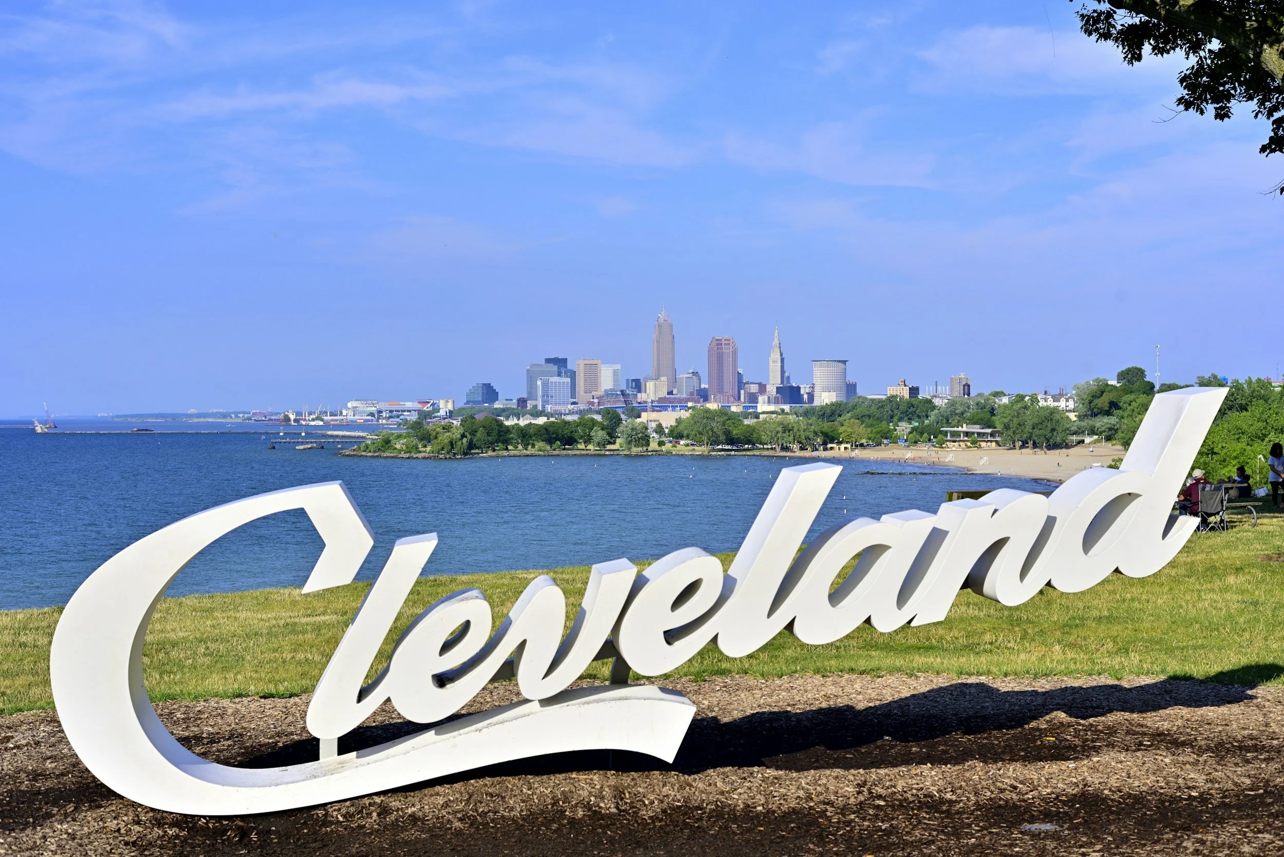 Cleveland city skyline viewed from a park with a large white sign that spells 'Cleveland' in the foreground, near a body of water and green grass.