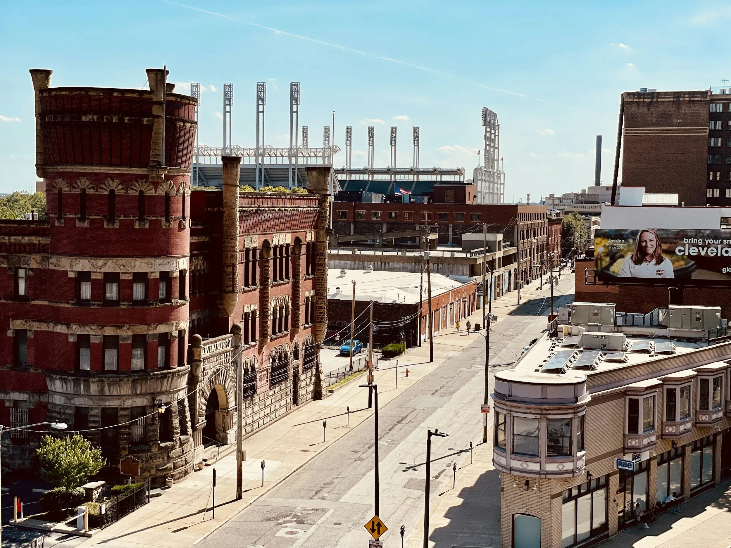 Cityscape of downtown Cleveland, featuring historic red brick buildings, modern stadium in the background, and a billboard with a smiling woman on it.