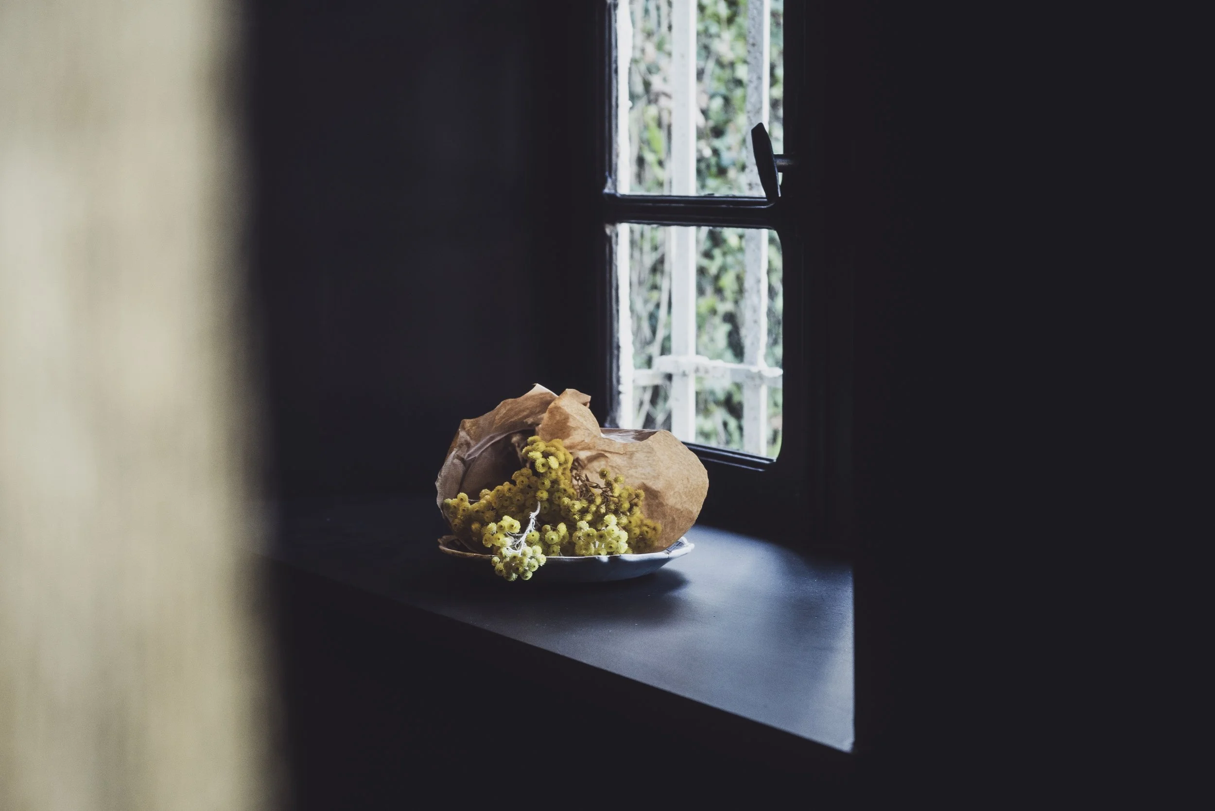 Un bouquet de petites fleurs jaunes dans un papier kraft posé sur une surface noire près d'une fenêtre ouverte.