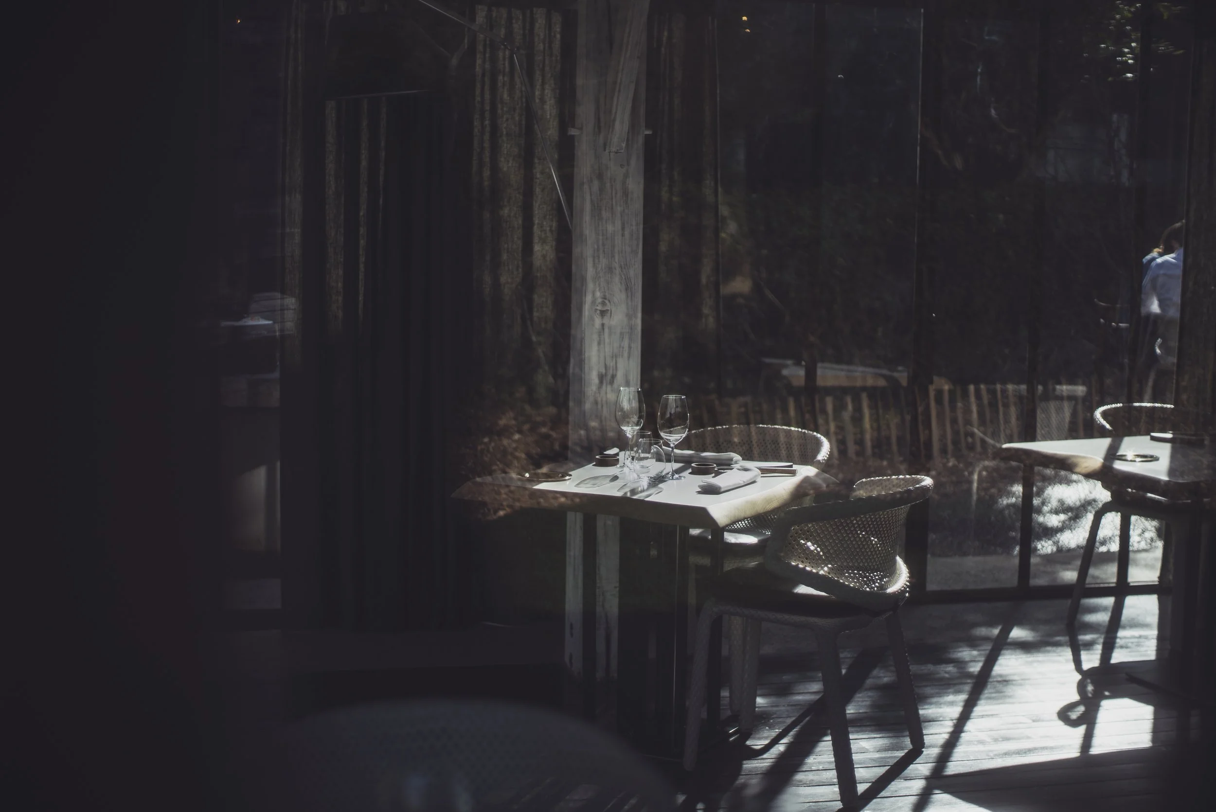 Une table du restaurant Le Moulin d'Alotz avec deux verres à vin, des assiettes, et des couverts, vue à travers une fenêtre avec reflection d'une jardin