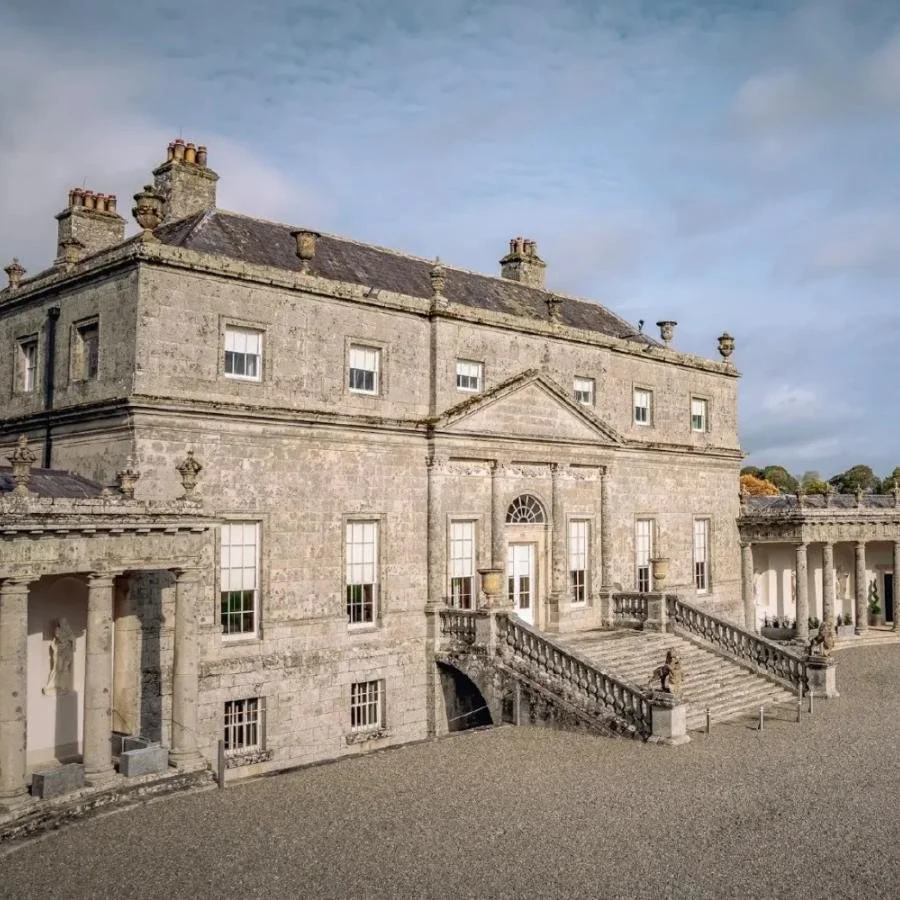 A large historic stone mansion with multiple chimneys, situated above a stone staircase and front steps, surrounded by a gravel courtyard and a colonnade of columns.