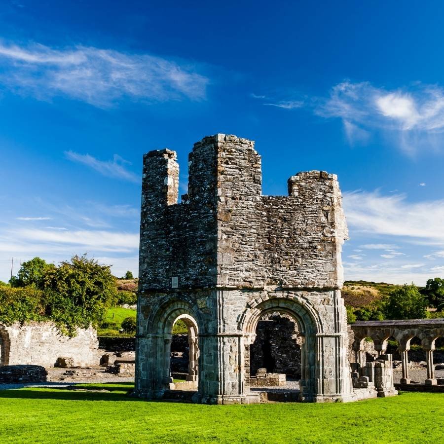 Ancient stone medieval tower with arches at its base, surrounded by green grass and ruins under a bright blue sky with wispy clouds.