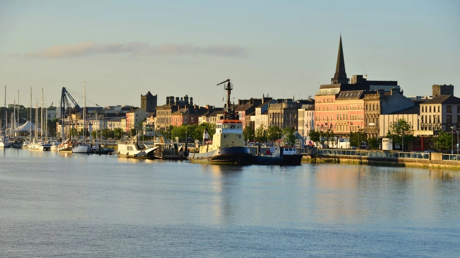 A harbor with boats docked along a waterfront promenade, with colorful buildings and a church with a tall spire in the background.