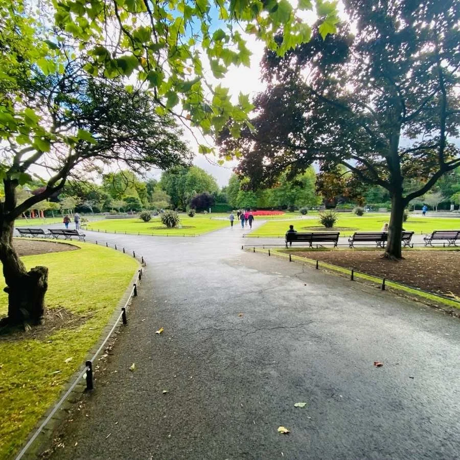 A park with green grass, trees, benches, and a paved walking path. Several people are walking and sitting, with a group in the distance.
