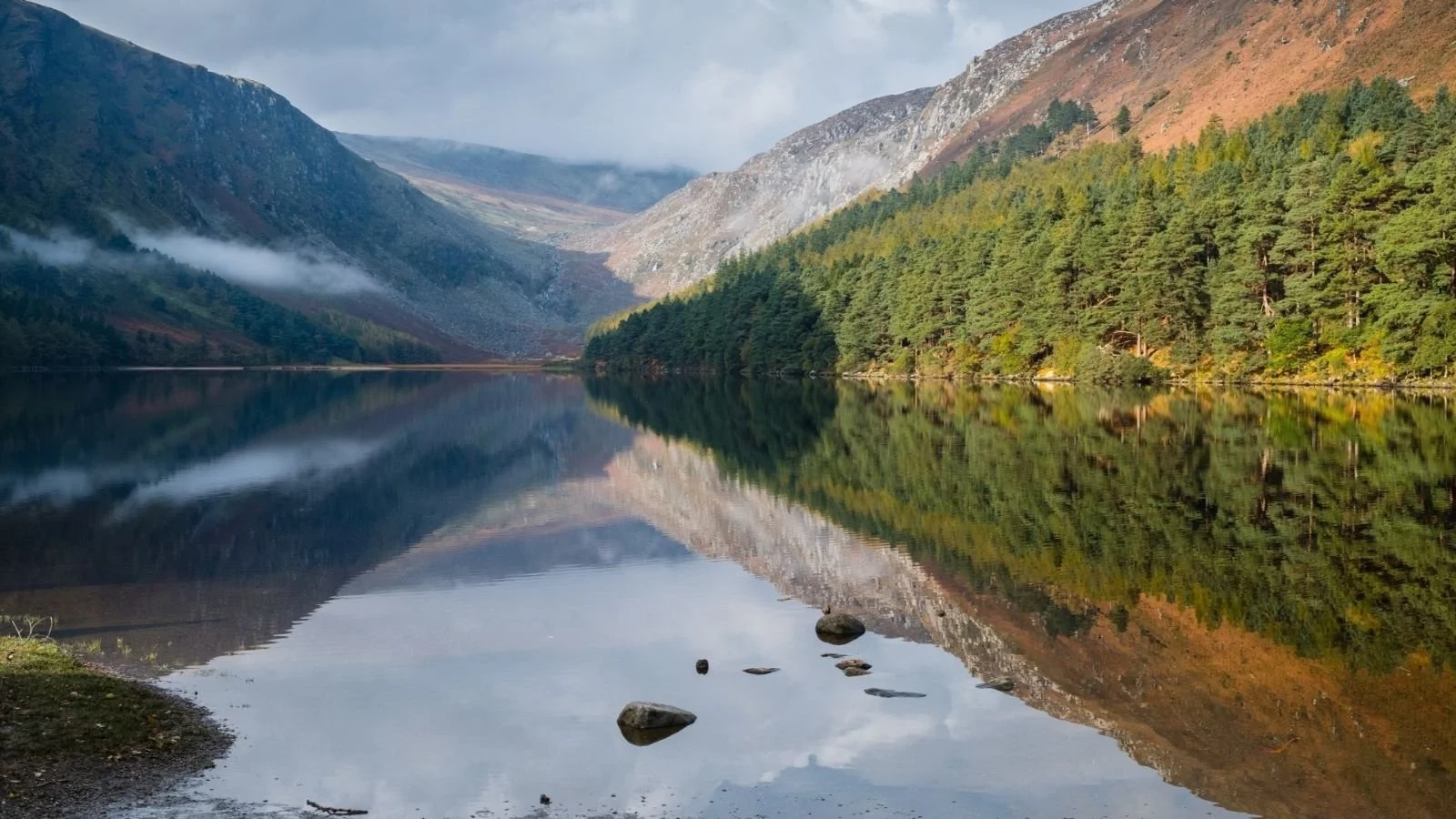 A calm lake reflecting green trees on a hillside and mountains in the background, with a partly cloudy sky.