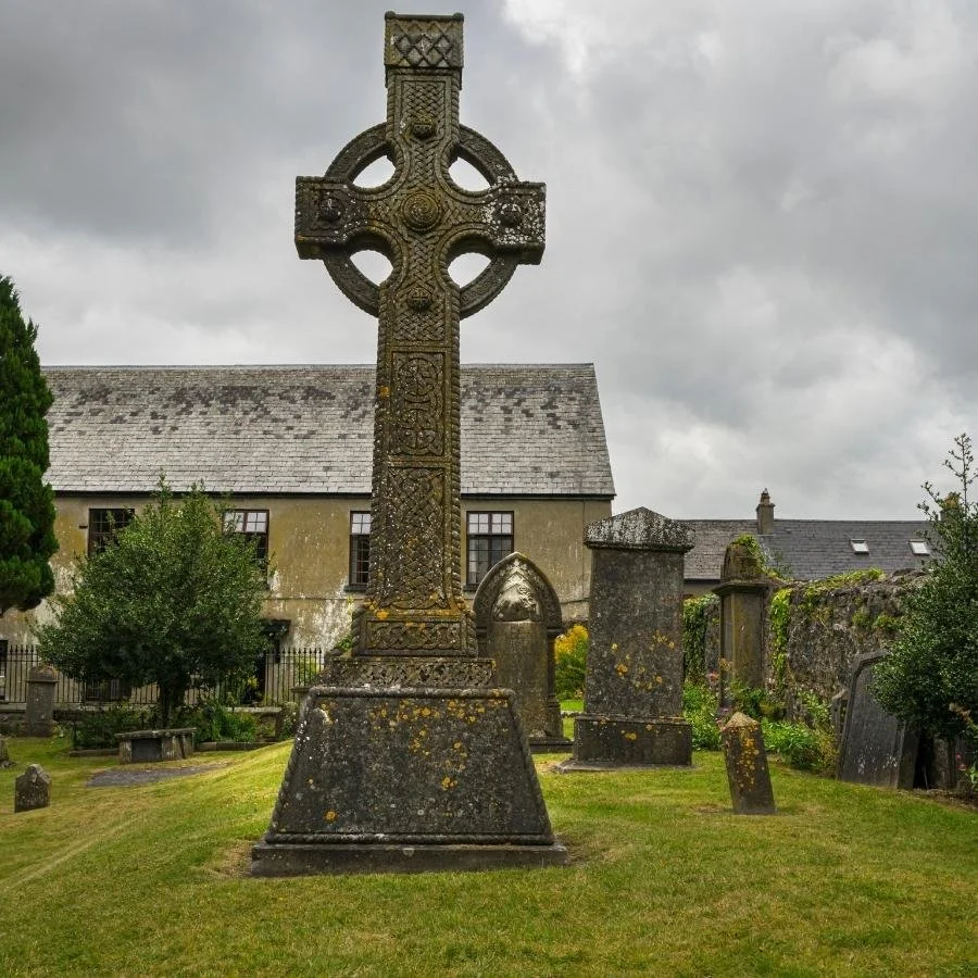 An old Celtic cross headstone in a historic graveyard with several other headstones and a building with a gray shingle roof in the background.