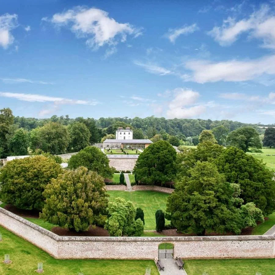 A lush green landscape with trees, grass, stone walls, and a white building in the distance under a partly cloudy sky.