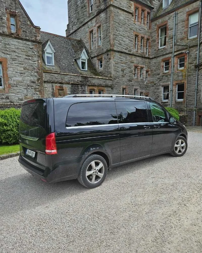 A black Mercedes-Benz van parked on a gravel driveway in front of a stone castle-like building with multiple windows and a roof with dormer windows.