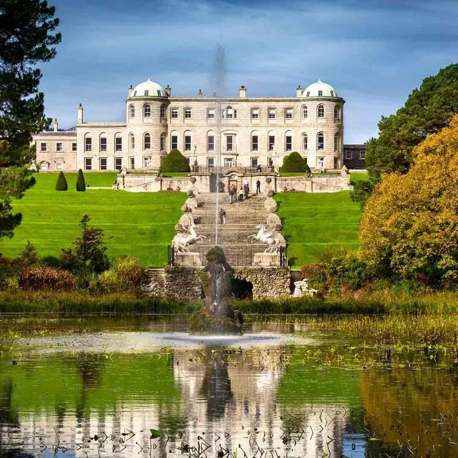 A large, historic white mansion with domed towers sits atop a green hill, overlooking a small pond with a fountain and statue in the foreground, surrounded by trees with fall foliage.