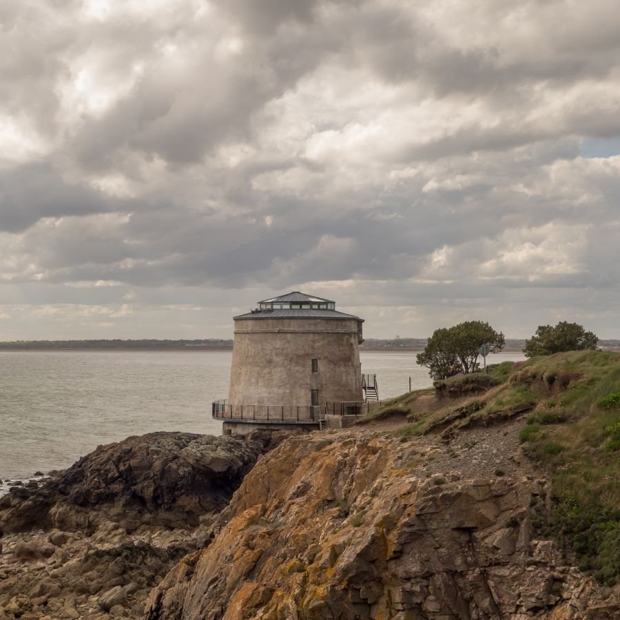 A historic stone lighthouse on a rocky cliff overlooking the water on a cloudy day.