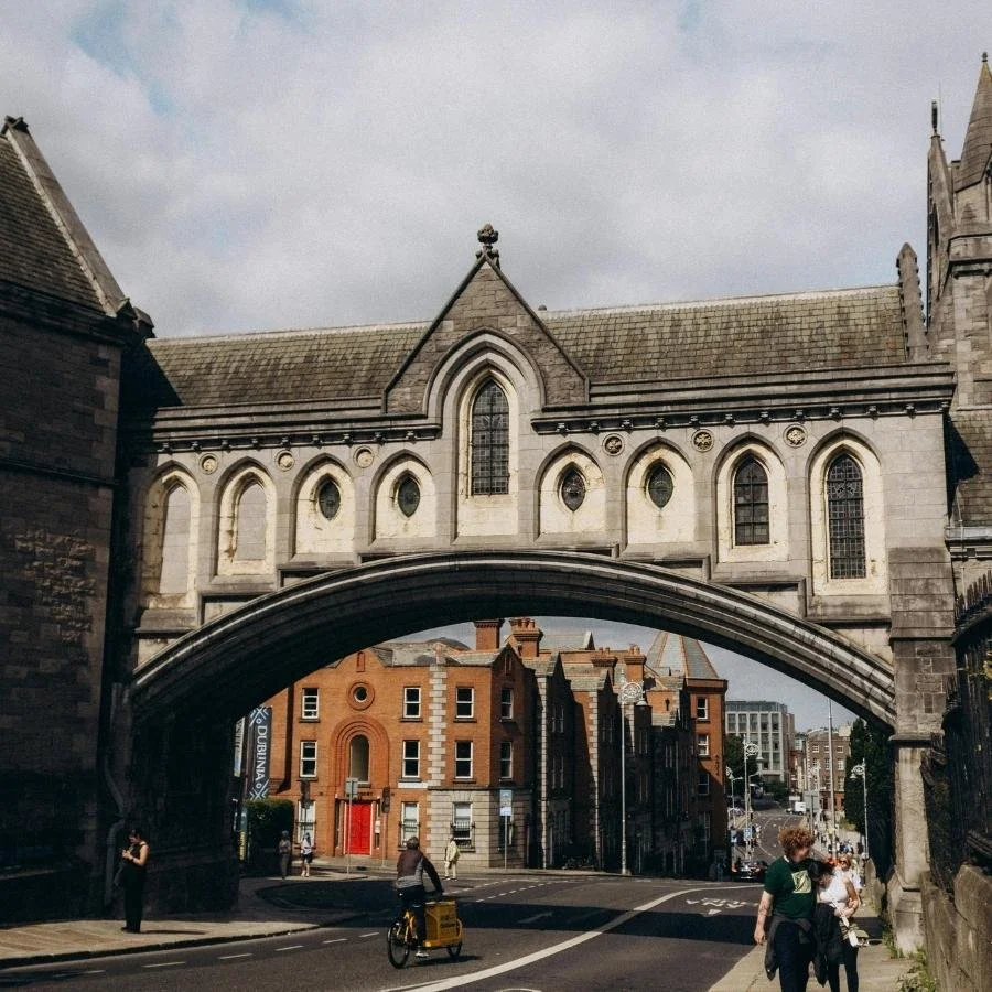 A historic stone building with arched windows and a bridge connecting two parts of the structure over a city street with pedestrians and a cyclist.