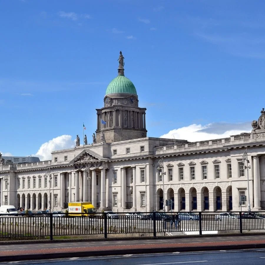A historic building with a green dome, columns, and statues on top, situated in a city with cars and a yellow delivery truck in front.