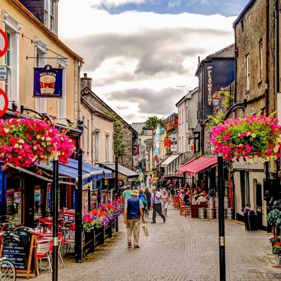A lively pedestrian street lined with cafes and shops, decorated with hanging flower baskets, with people walking and sitting outside eateries under a cloudy sky.