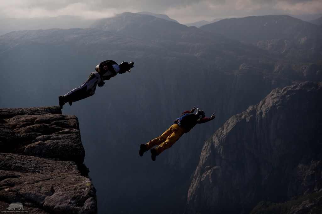 Two skydivers in mid-air jumping from a cliff over a deep canyon with mountains in the background.