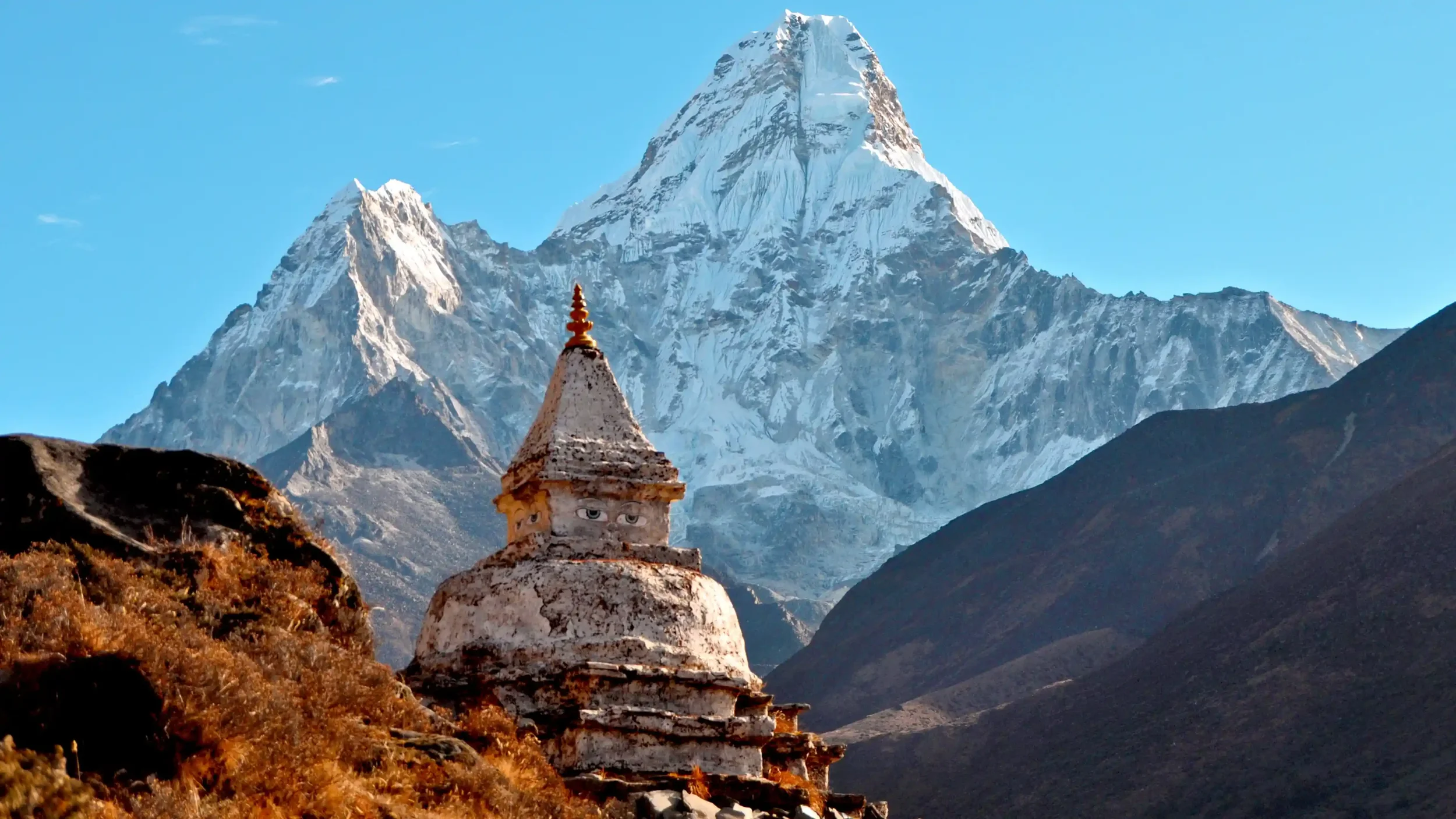 Snow-capped mountains with a small, ancient temple with eyes painted on its front, in a mountainous landscape with clear blue sky.