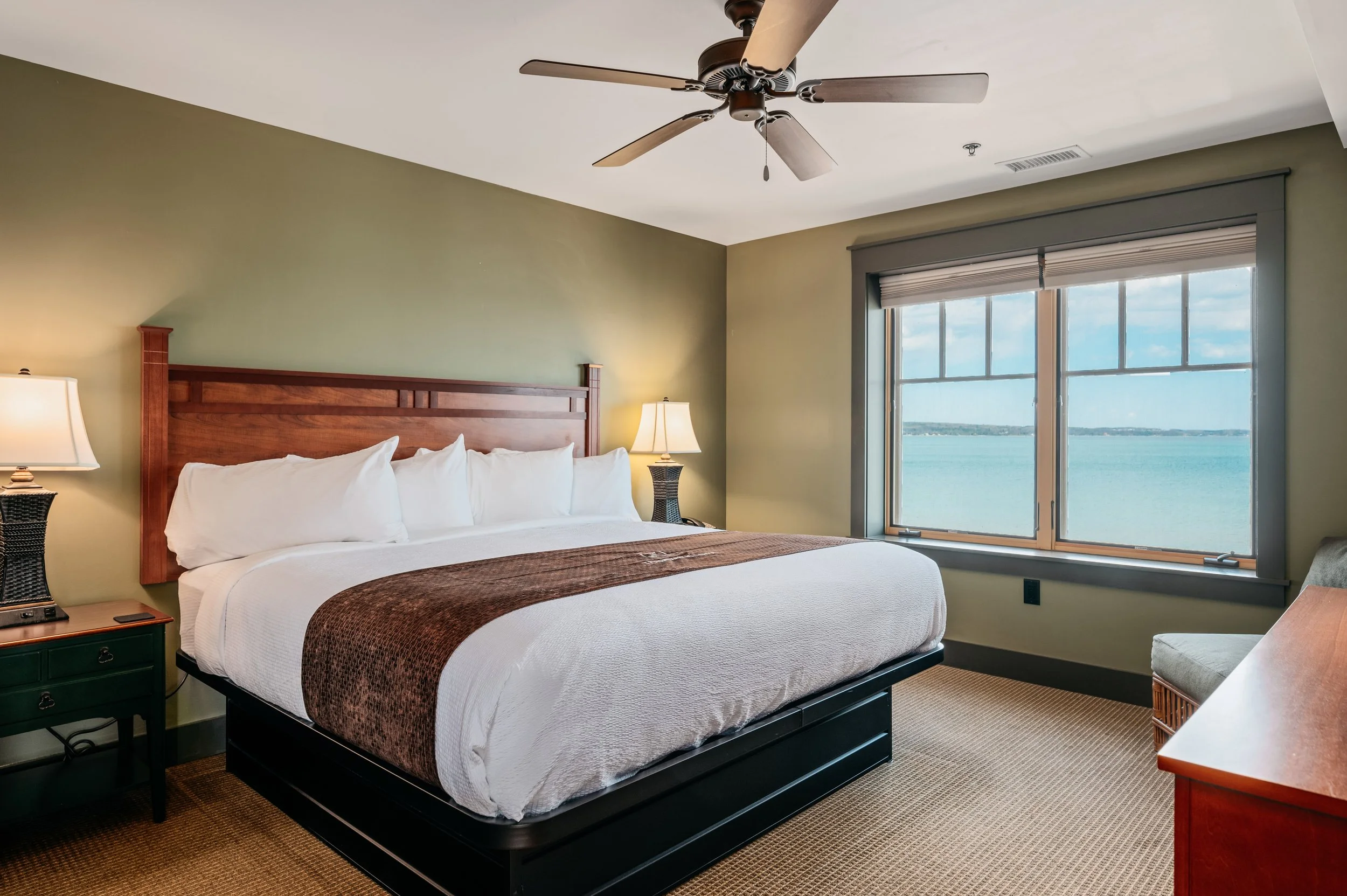 A hotel room with a large bed, wooden headboard, white bedding, brown throw blanket, two bedside lamps, a window showing a view of water and sky, ceiling fan, and nightstand.