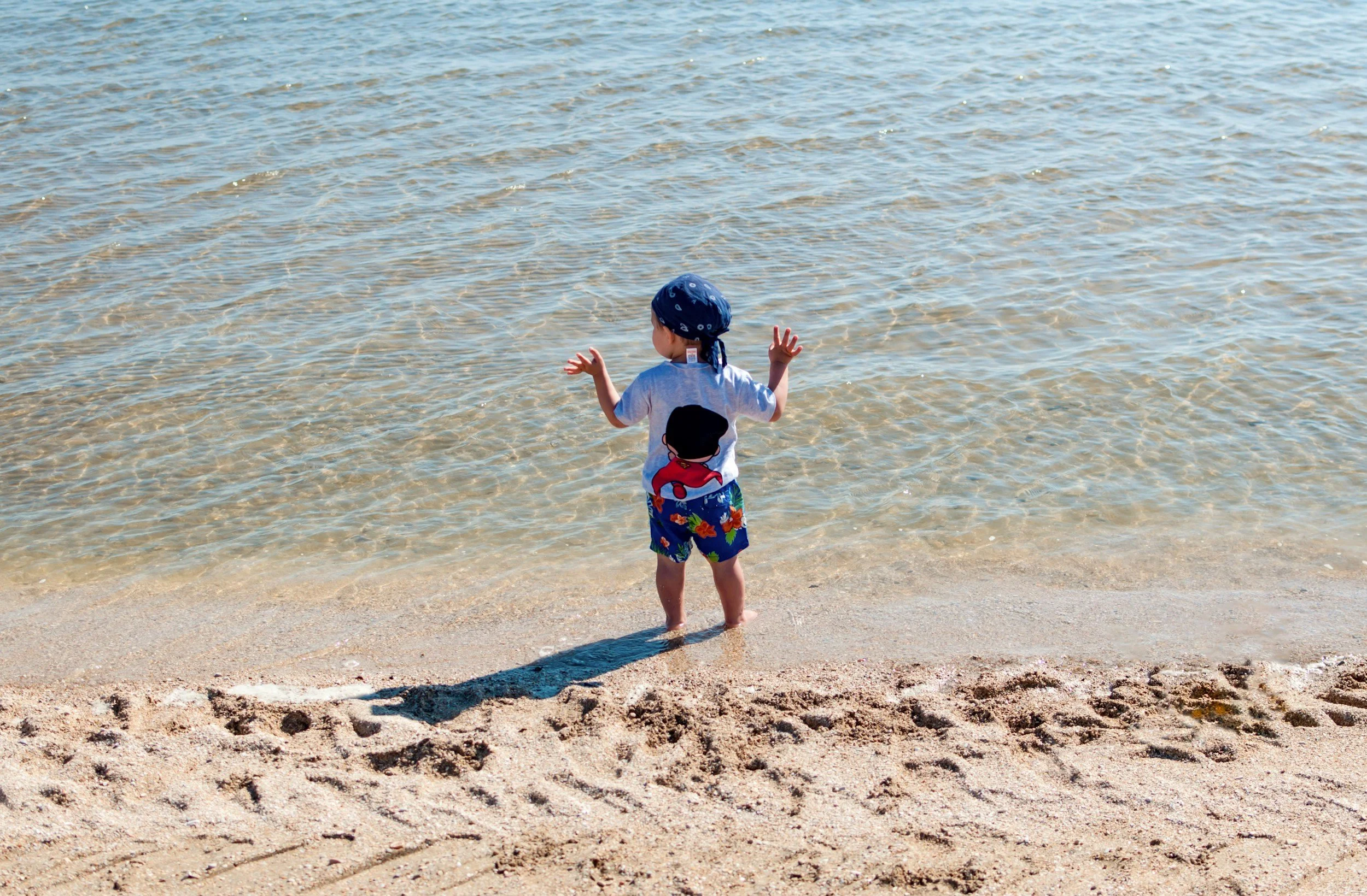 A young child standing at the water's edge on a sandy beach, facing the ocean with arms raised, wearing a blue bandana, a white T-shirt with a cartoon character on the back, and colorful swimming trunks.