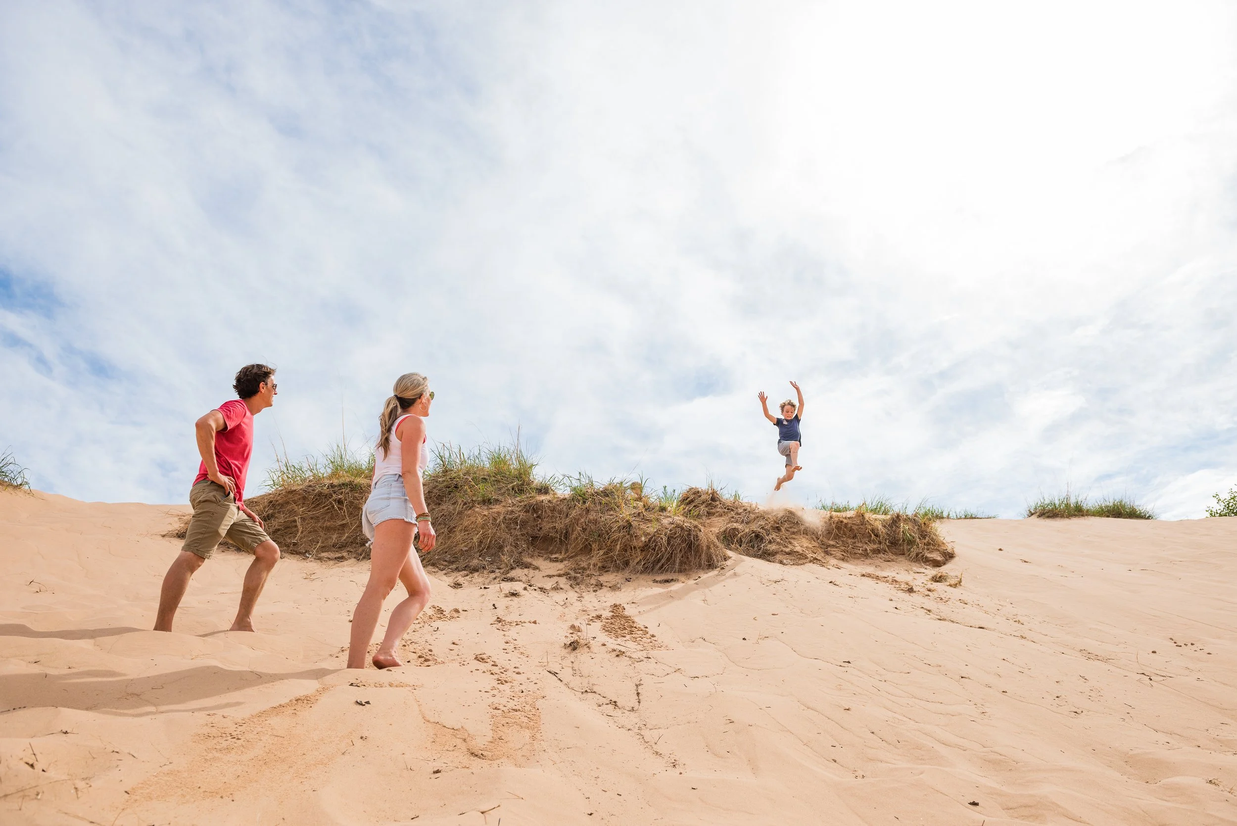 A family of three at the beach, with two adults watching as a child jumps off a sand dune.