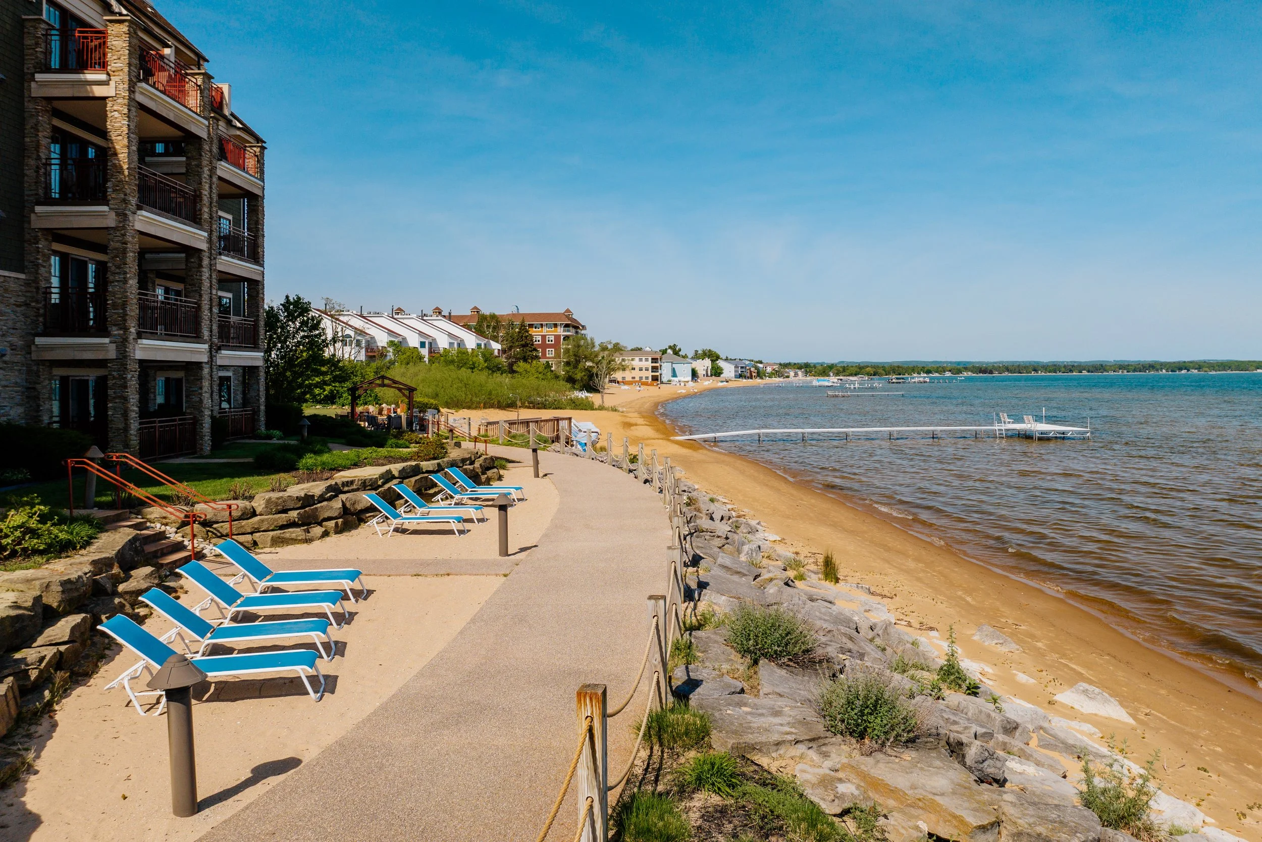 A lakeside scene with a paved walkway, blue lounge chairs, and a building on the left side. The sandy beach extends along the shore, with boats docked in the water. There are houses and trees in the background under a clear blue sky.