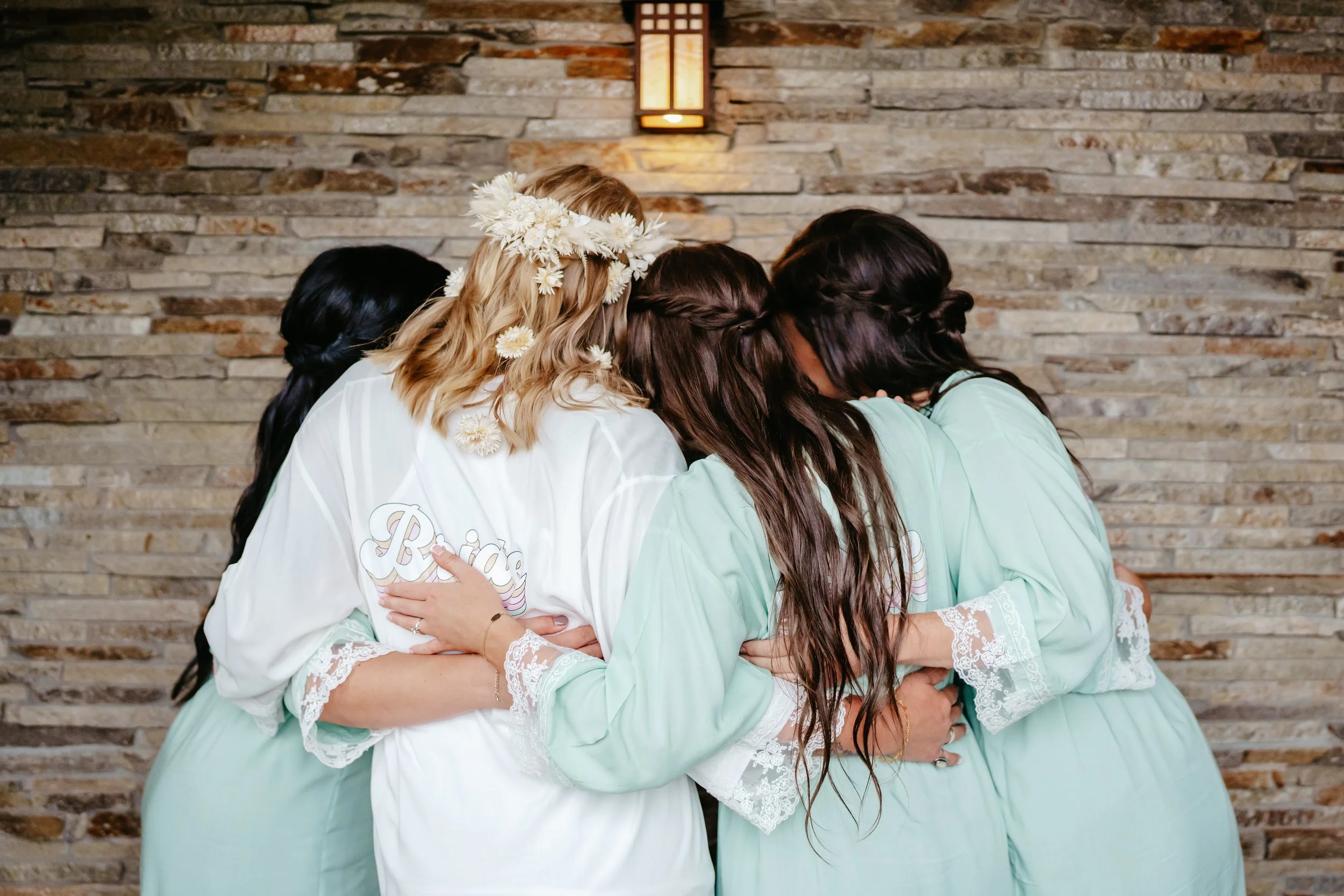 Group of women in pastel robes hugging each other in front of a stone wall, with one woman wearing a flower crown and a robe that says 'Bride'.