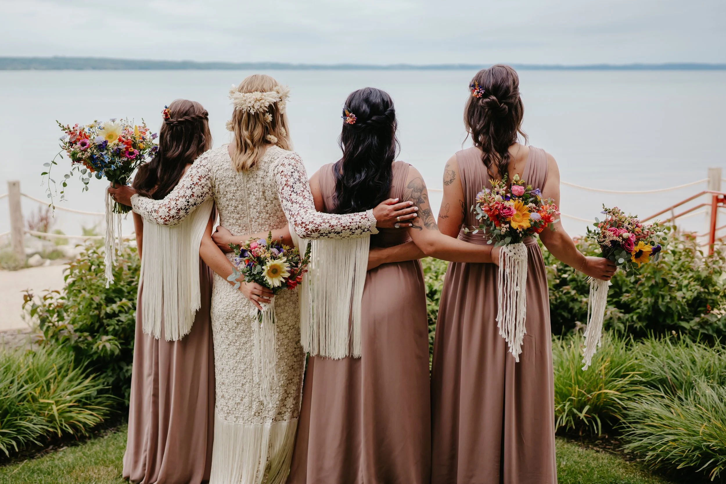 Group of women in boho-chic dresses holding bouquets, standing by the water at a beachside location.