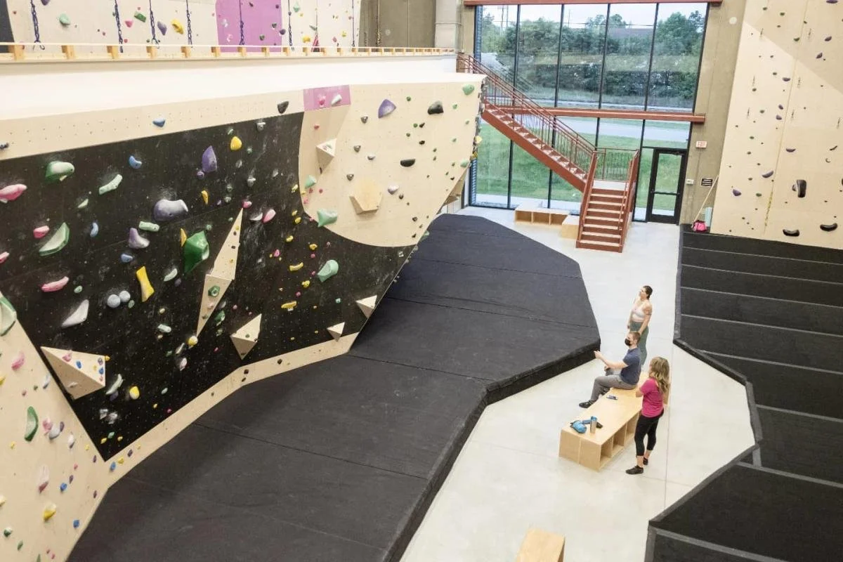 Indoor rock climbing gym with artificial climbing walls, including a large overhang section, and a sitting area with three people inside.