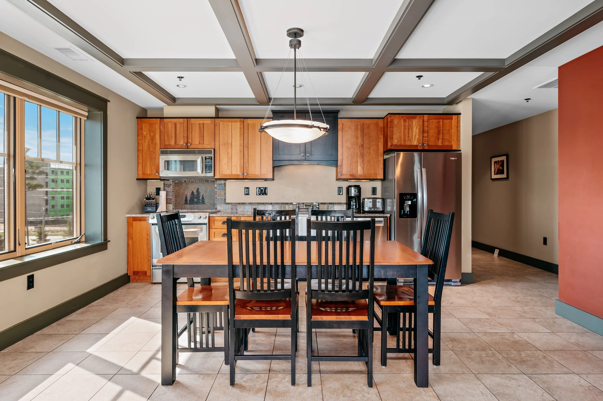 Modern kitchen with wooden cabinets, stainless steel appliances, a dining table with six black chairs, and a large window letting in natural light.