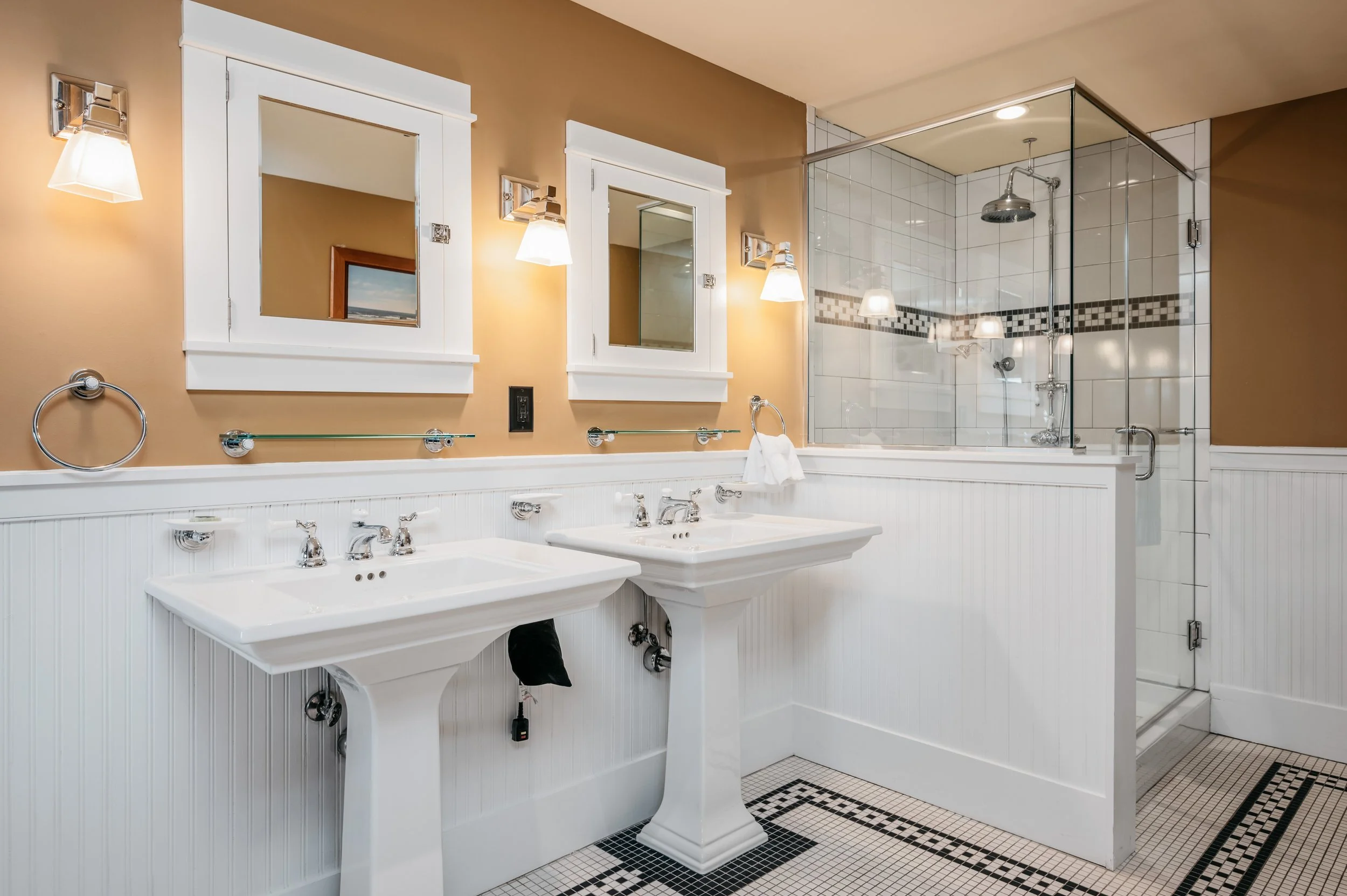 Bathroom with two white vintage pedestal sinks with mirrors and wall-mounted lights, a glass-enclosed shower with white tiles and black accent trim, tan walls, and a black and white tiled floor.