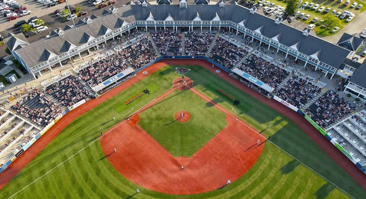 Aerial view of a baseball stadium filled with spectators and players, with parking lots surrounding the venue on a sunny day.