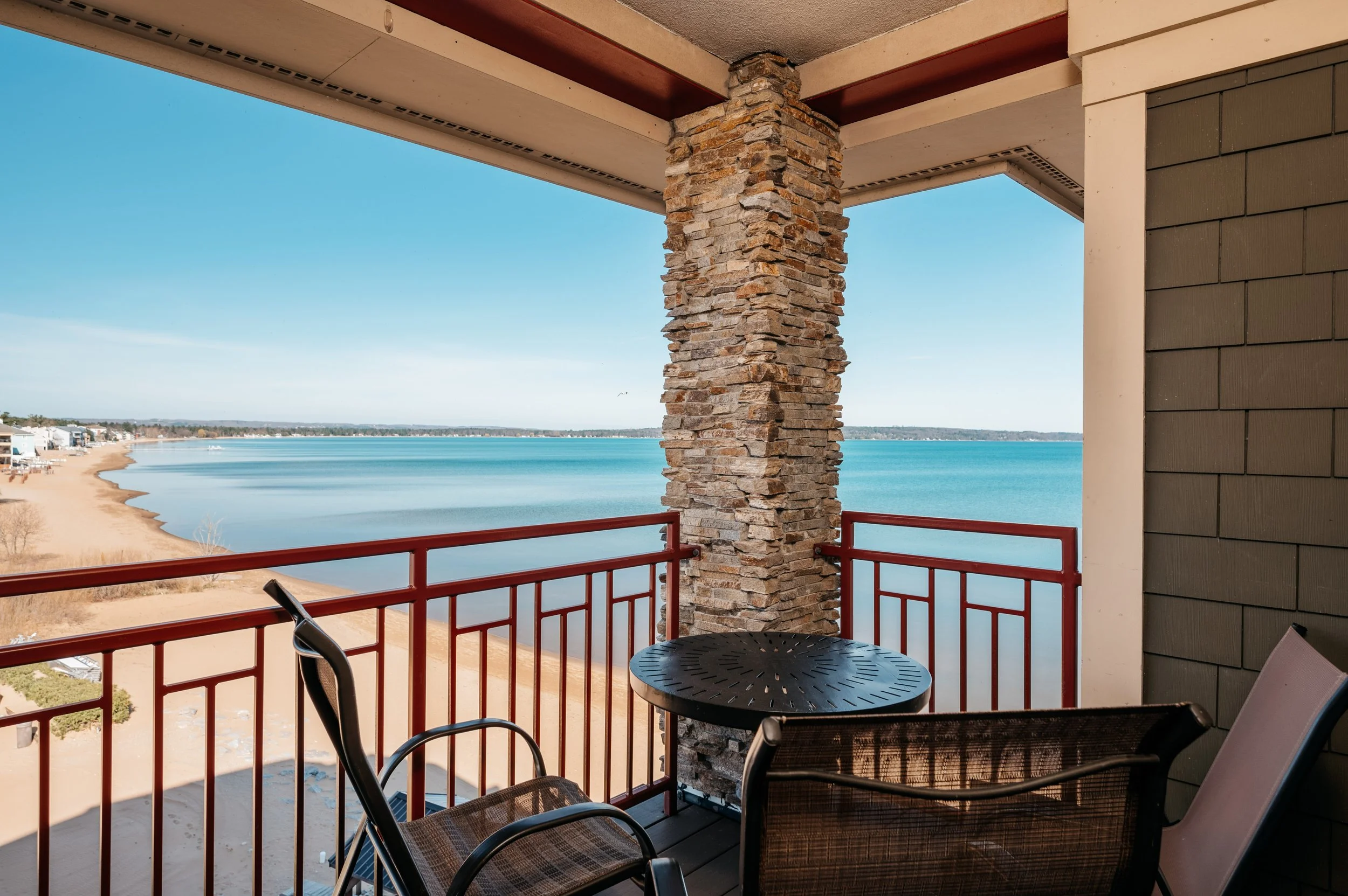 Balcony view overlooking a lake or ocean, with sandy shoreline and houses in the distance, featuring red metal railing, stone column, and outdoor seating.