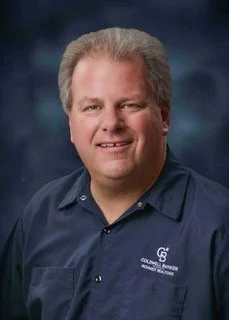 A man with light hair wearing a navy blue collared shirt against a dark background.