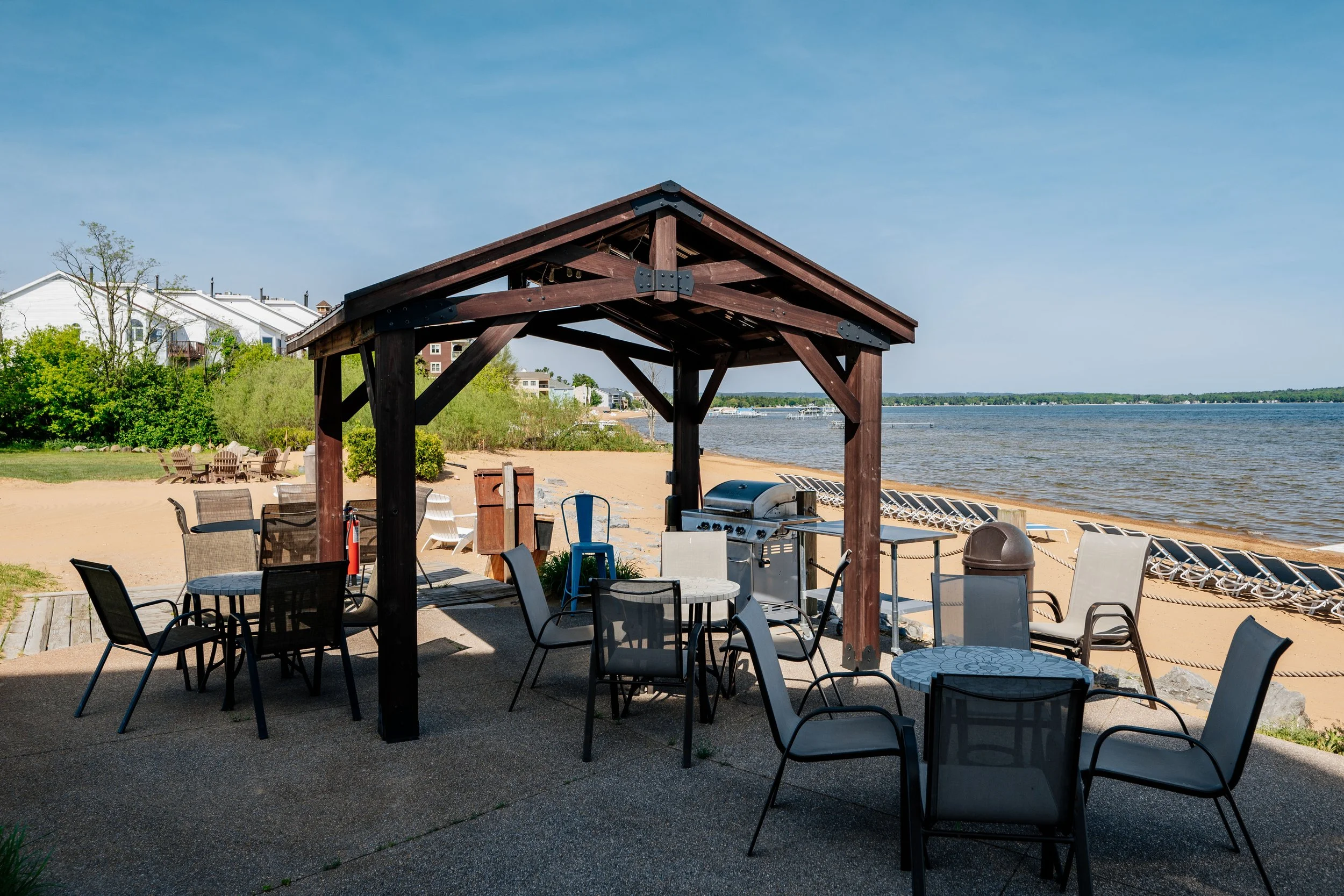 Beachside patio with tables, chairs, and a covered grill area, overlooking a sandy beach and a body of water.