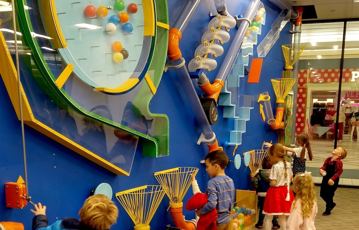 Children playing a game at an arcade with a large Rube Goldberg machine involving colorful tubes, pulleys, and balls.