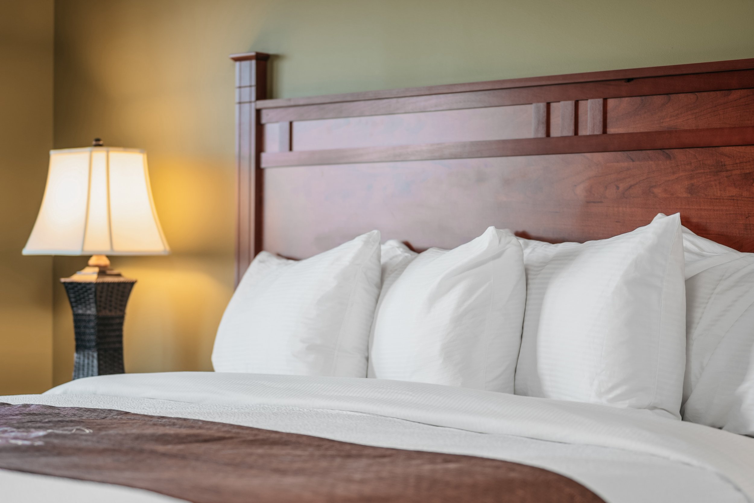 Freshly made bed with white pillows and beige bedding, bedside lamp with a dark woven base, wooden headboard, and pale yellow wall.