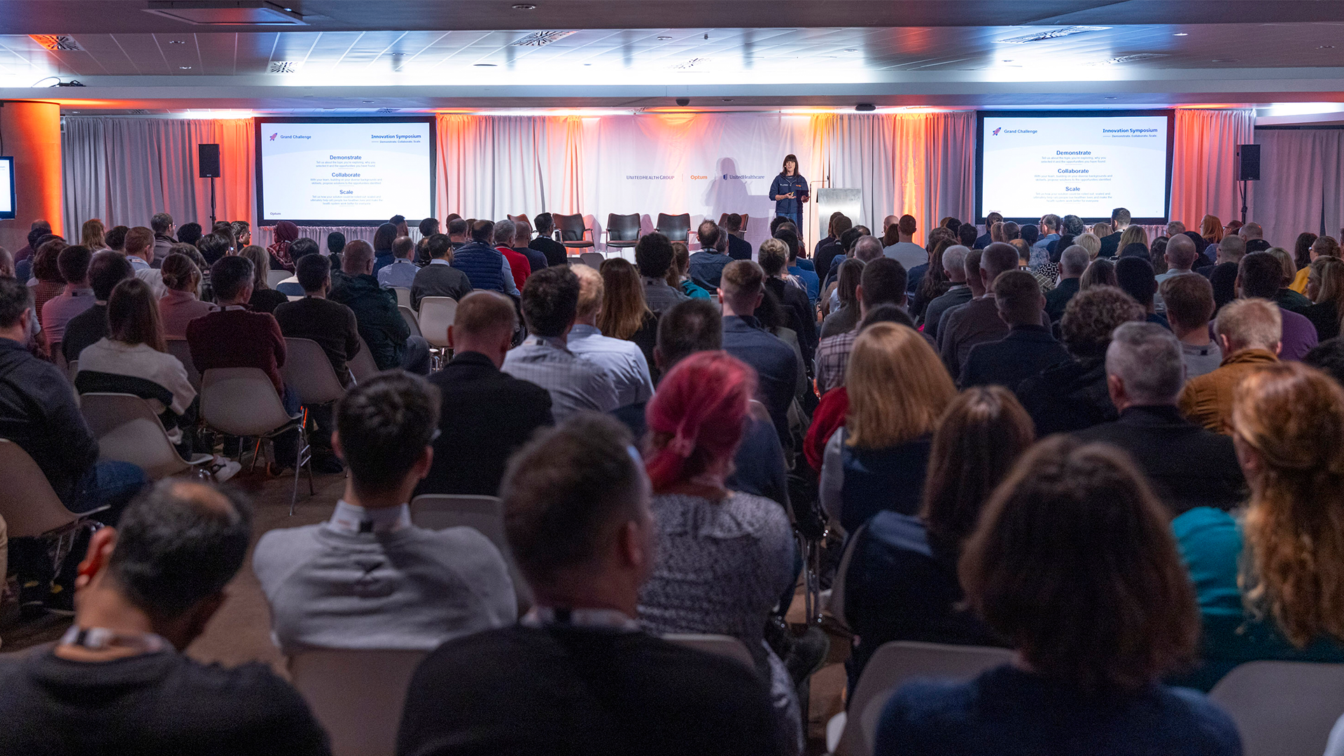 A large conference room filled with a diverse audience listening to a speaker on stage. The stage has two large screens displaying presentation slides about demonstrating, collaborating, and scaling. The room has white curtains and orange lighting accents.
