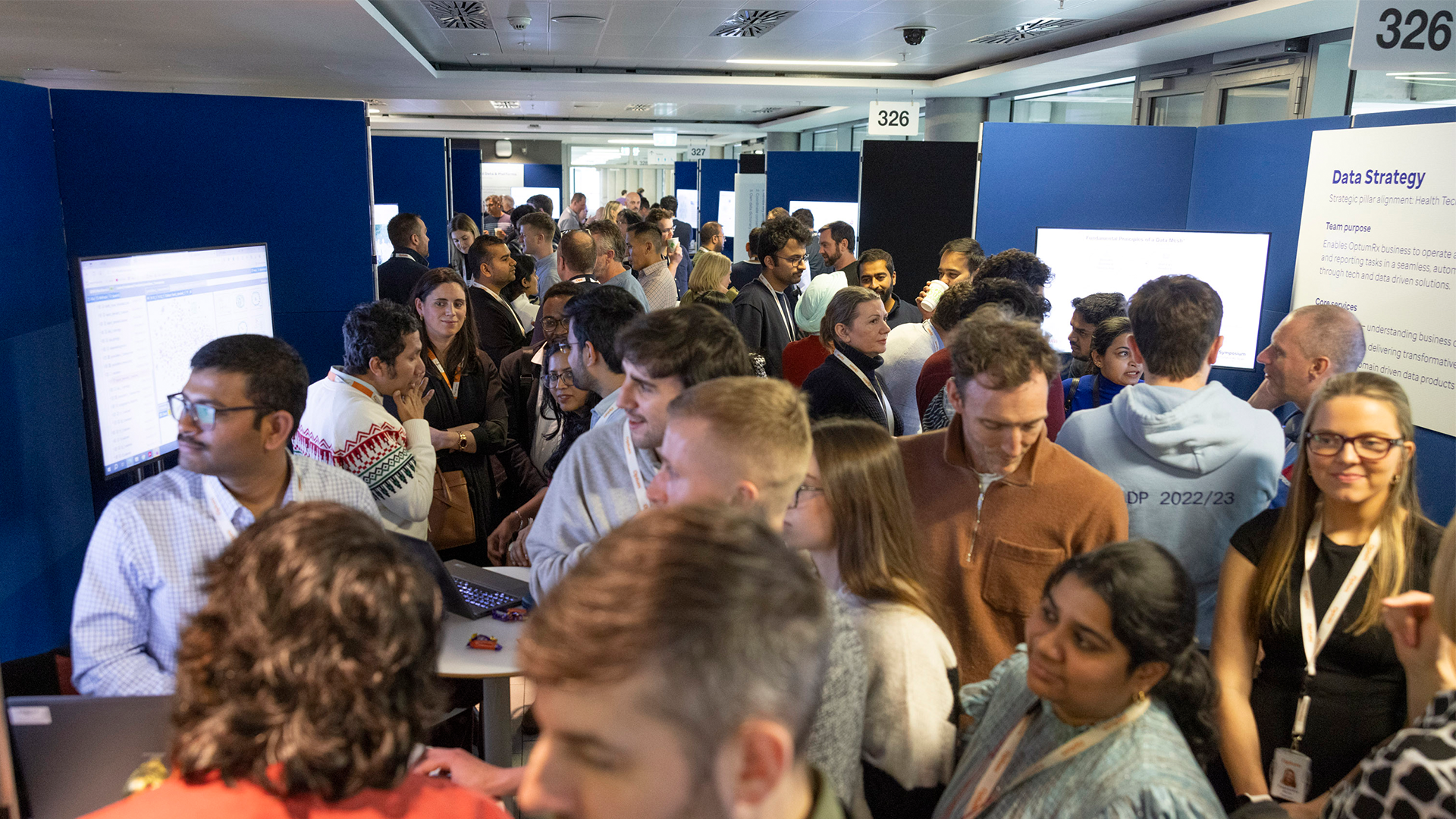 Crowded conference exhibit hall with numerous people engaging in conversations near blue exhibition panels, some standing in front of screens displaying data and information.