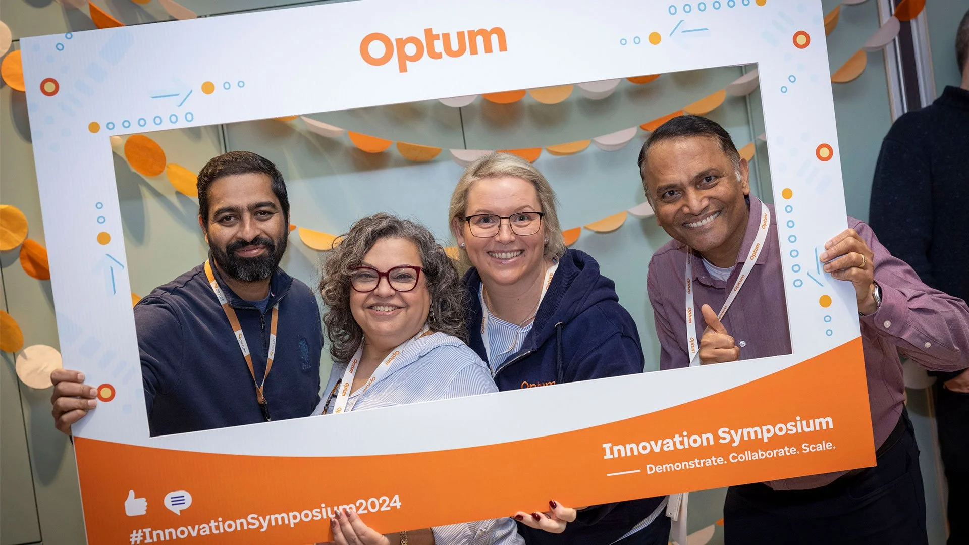 Four diverse people smiling and posing behind a large orange and white frame at the Optum Innovation Symposium 2024 event.
