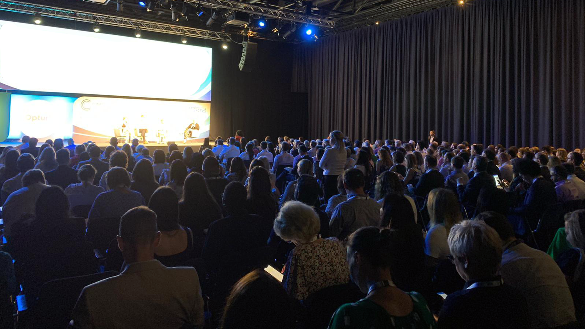 Large auditorium space filled with people and four speakers on a stage