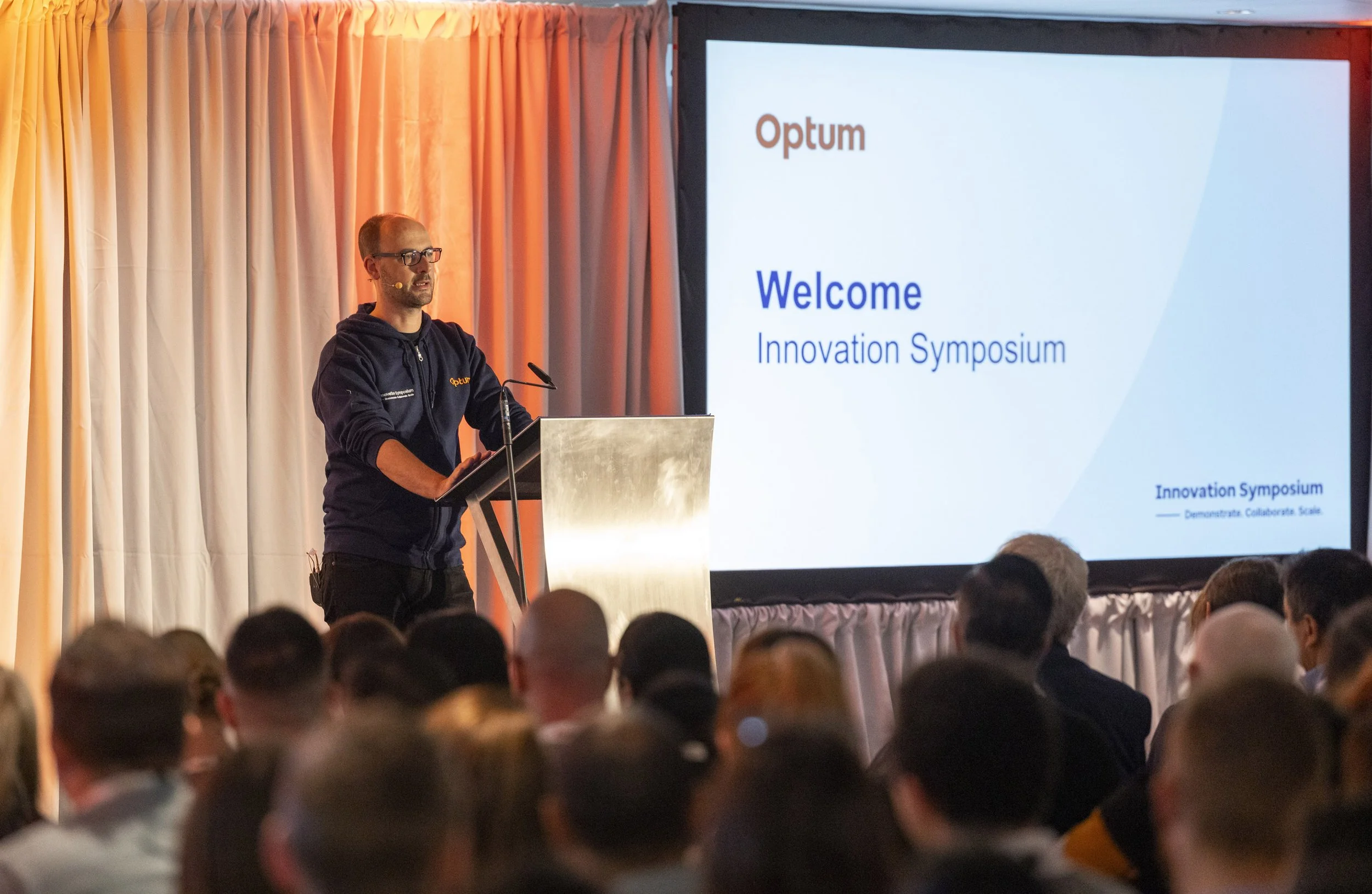A man is speaking at a podium during an Innovation Symposium, with a large screen behind him displaying the words 'Optum' and 'Welcome, Innovation Symposium.' The audience is seated in front of him, and the room is decorated with curtains.
