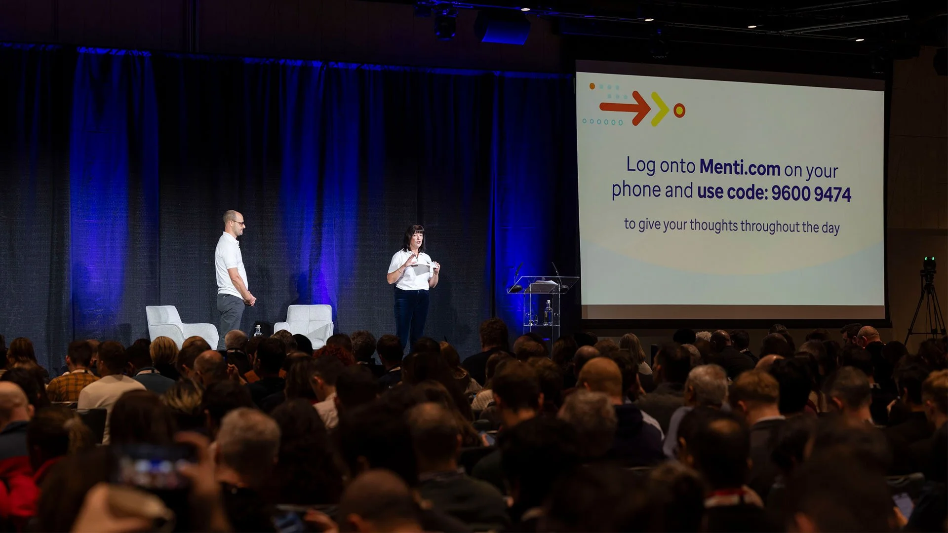 A conference presentation with two speakers on stage, one man and one woman, with a large screen displaying instructions to log onto Menti.com and use a specific code, while the audience watches.