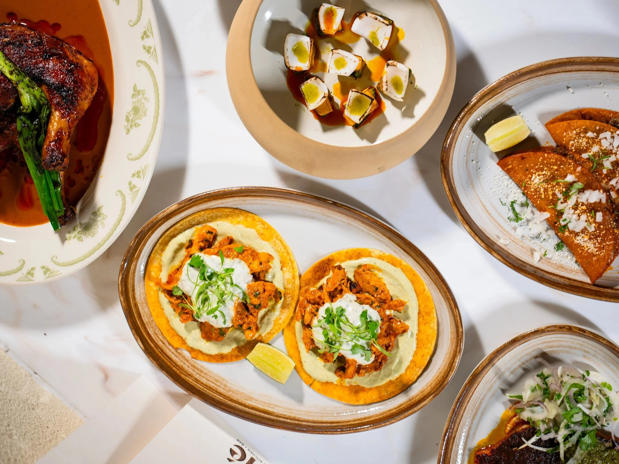 Assorted Indian dishes including two plates of chaat topped with cilantro, a bowl of samosas, and other traditional items on a white surface.