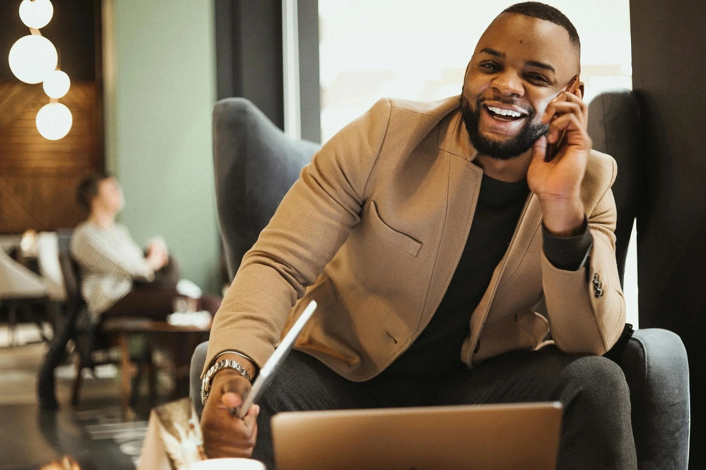 A smiling man with a beard wearing a beige blazer, sitting in a lounge, talking on a cellphone, with a laptop in front of him.