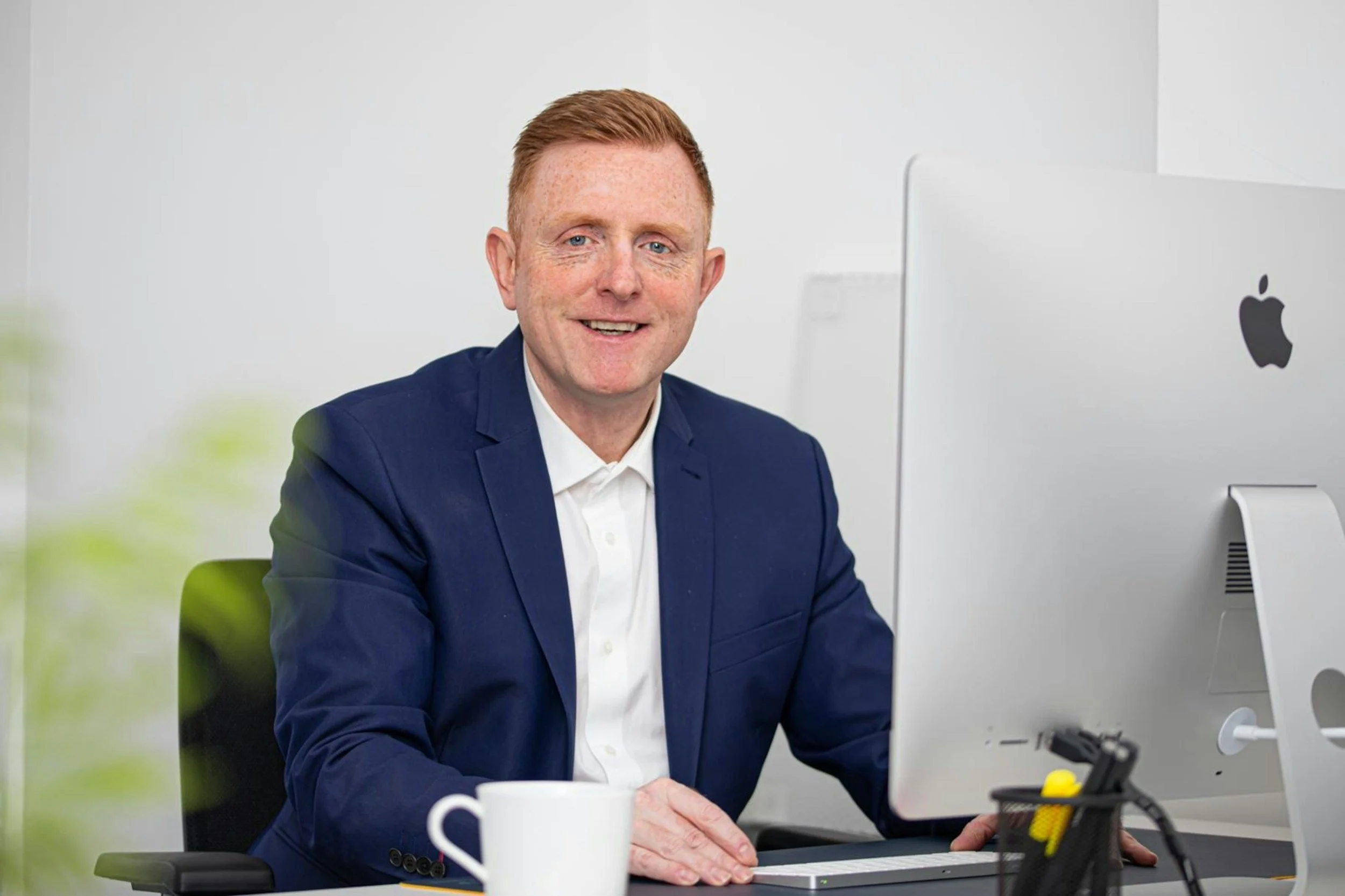 A man in a blue suit sitting at a desk with a white computer, a cup, and some office supplies, smiling at the camera in an office setting.