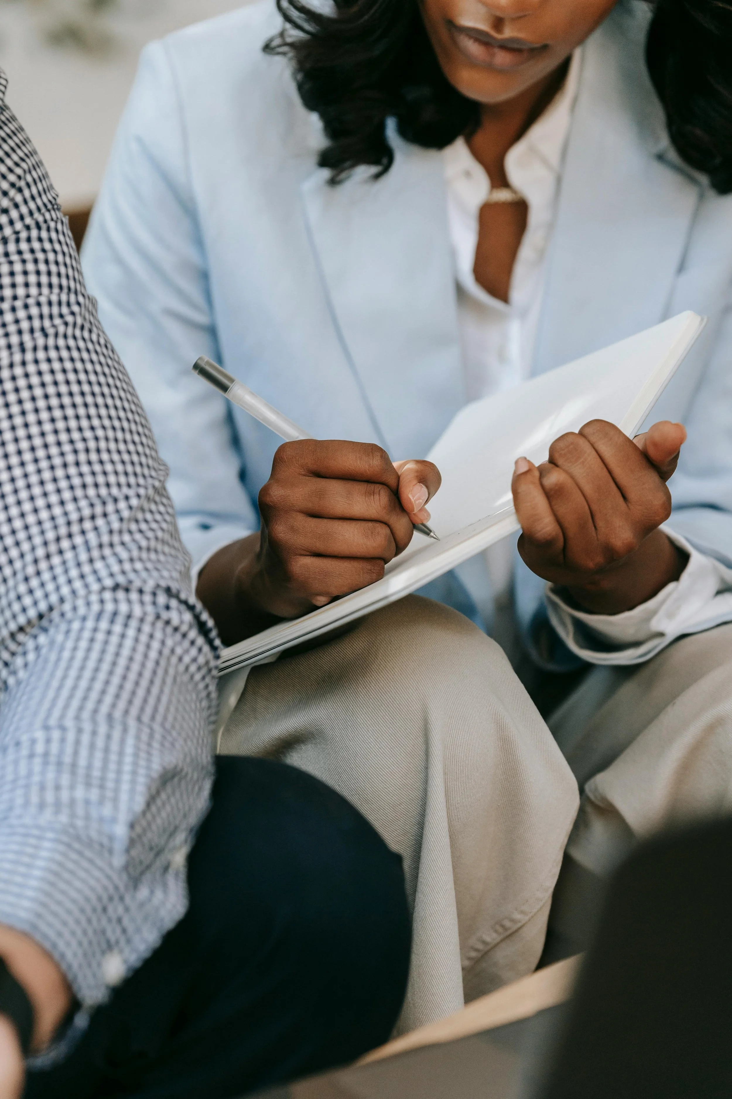 A person in a white blazer writing in a notebook with a pen, sitting close to another person in a checkered shirt.