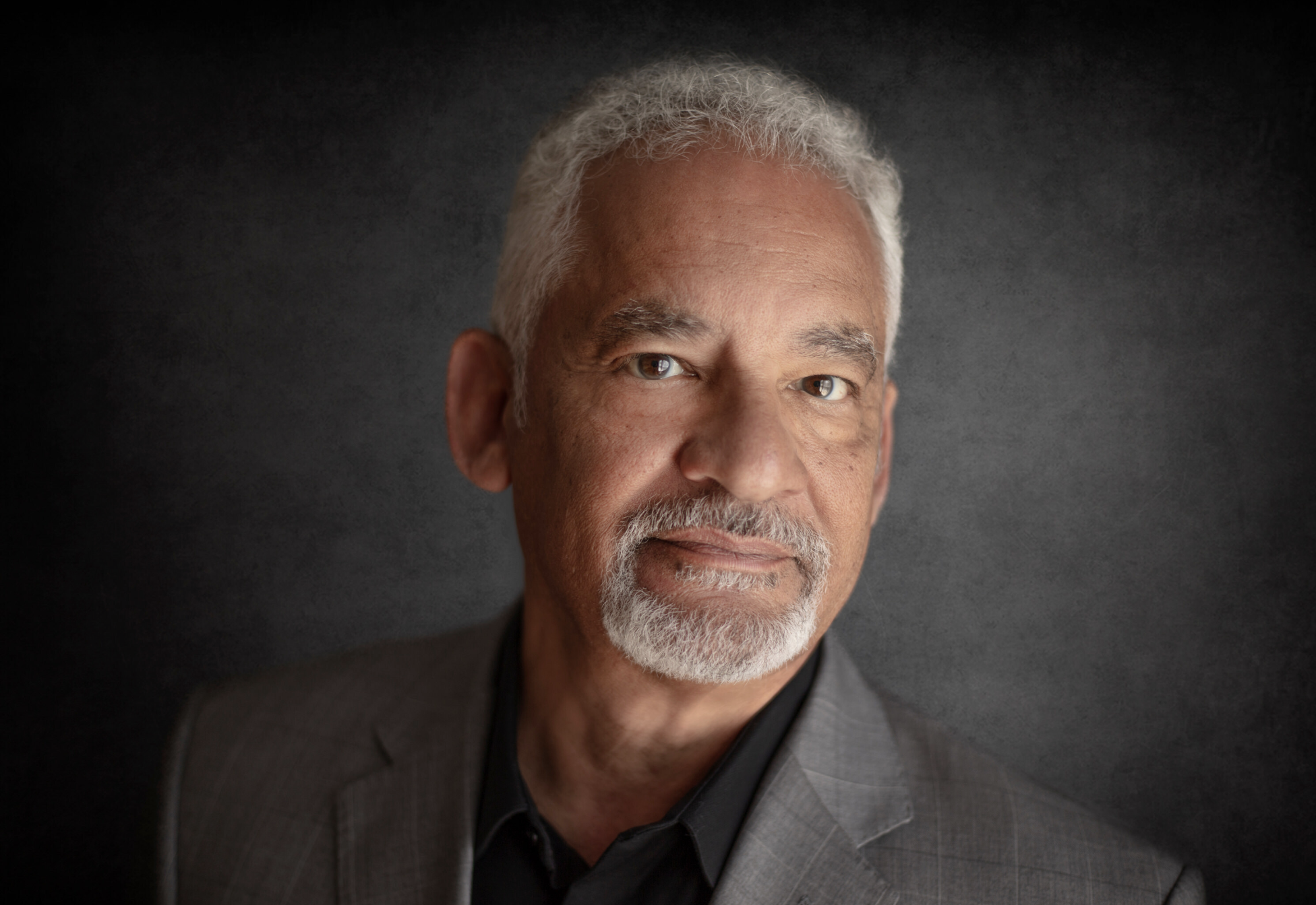 Close-up portrait of an older man with gray hair and beard, wearing a dark suit and black shirt, against a dark textured background.