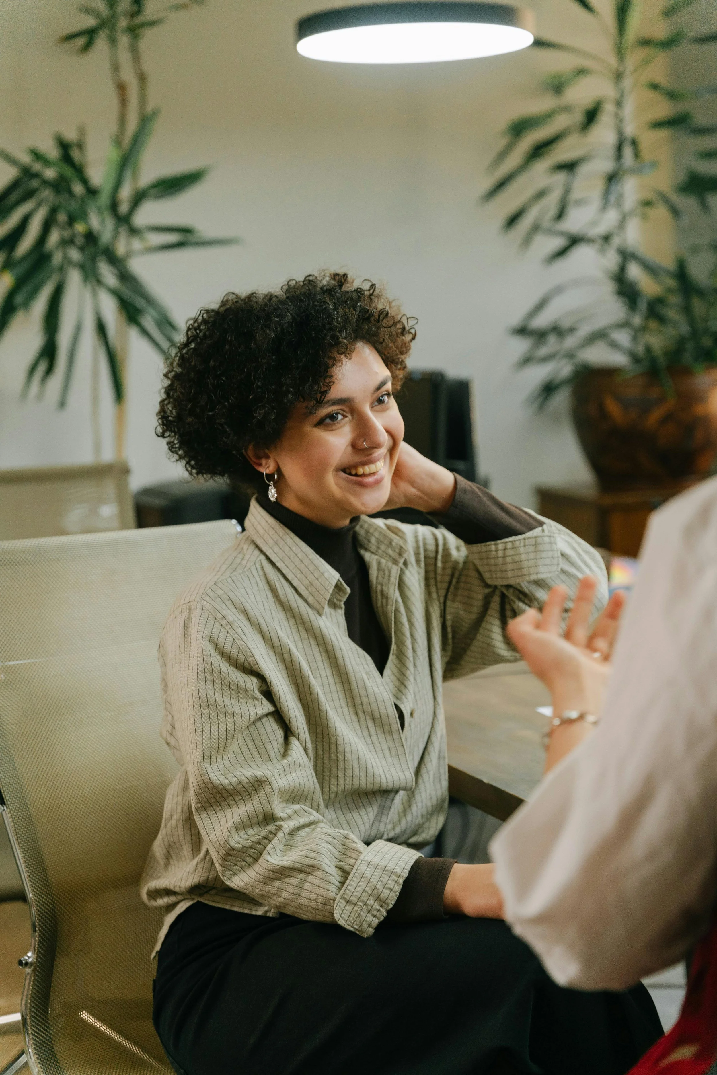 A woman with curly hair and earrings smiling as she listens to another person in a conversation in an indoor setting.